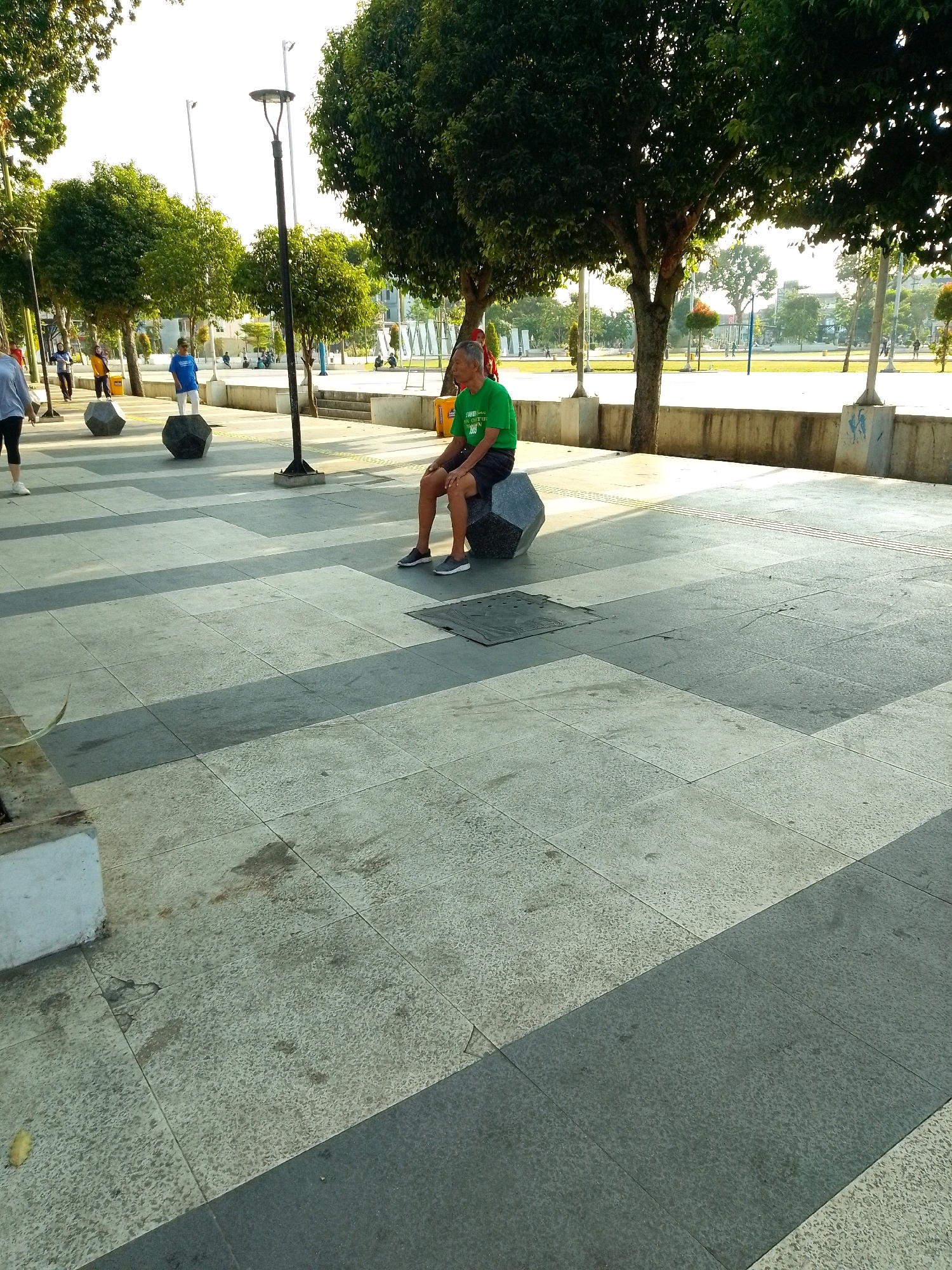 A man in a green t-shirt and black shorts is sitting on a large, dark, faceted stone bench in an outdoor paved area. The man is facing left, with his legs extended forward and his arms resting on his thighs. He is wearing dark grey sneakers. The paving stones are a mix of light grey and dark grey squares, laid out in a geometric pattern. In the background, there are several trees with green foliage, streetlights, and other people walking or sitting. There are also some concrete structures and a wall in the background. The lighting suggests it is daytime, possibly late afternoon, with sunlight casting shadows. The scene appears to be a public park or recreational area. This area is likely the Alun-Alun Jember, a popular public square in Jember, East Java, Indonesia, known for its green spaces and paved walking paths.