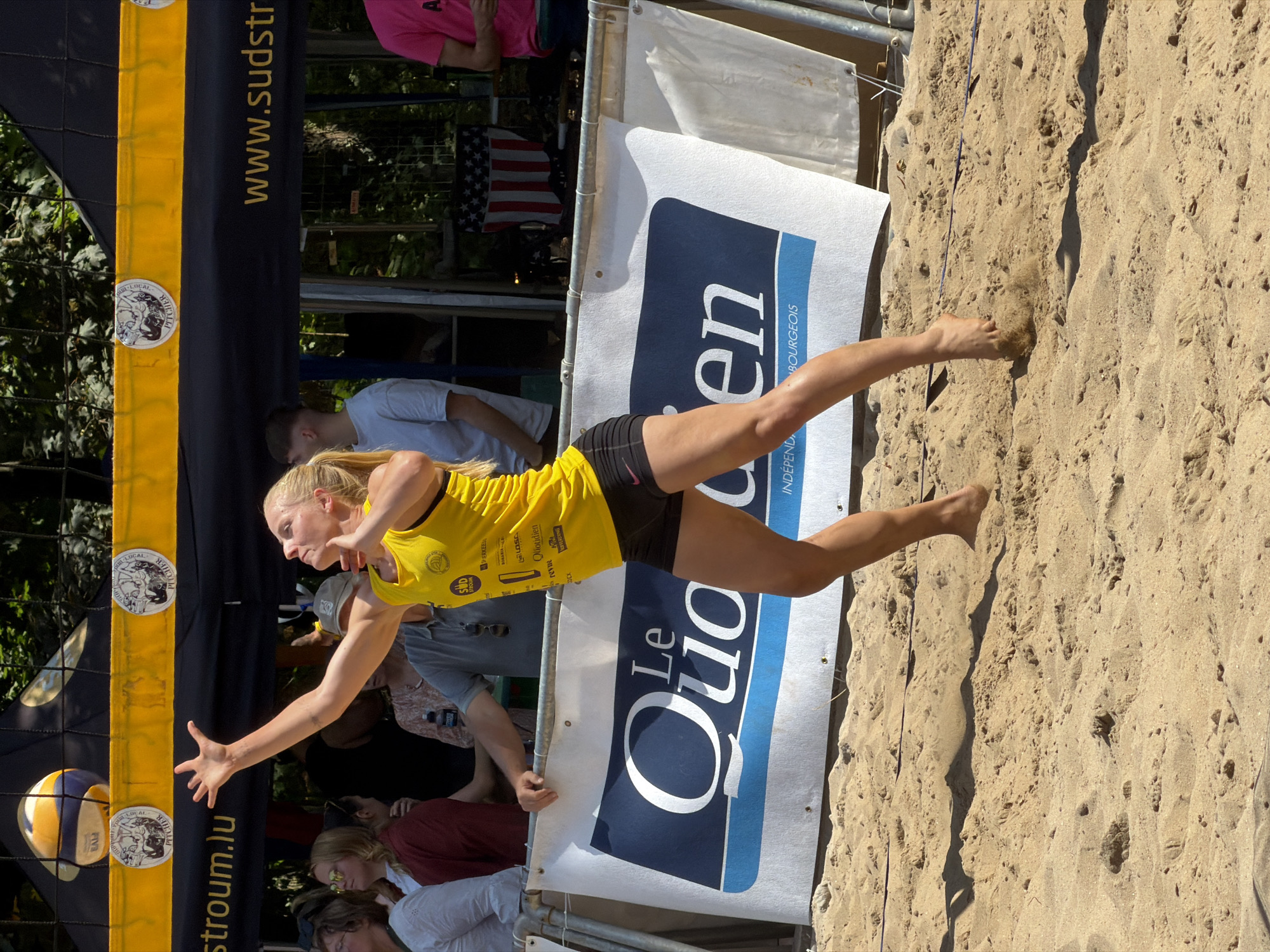 An outdoor, daytime, medium shot captures a blonde female volleyball player in mid-action on a sandy court. She is wearing a yellow tank top and black shorts, her left arm extended upwards as if to serve or set the ball, which is visible just above the net in the upper left quadrant of the image. Her focus is directed towards the ball. In the background, a dark blue tent with yellow trim and various logos is present. Text on the tent includes "stroum.lu" and "www.sudstr". A large white banner with the text "Le Quo en" in blue and white lettering is prominently displayed behind the player, with smaller text below reading "INDEPENDANT LUXEMBOURGEOIS". Other spectators are partially visible behind the banner and tent. The court is composed of fine, golden sand, with shadows cast by the players and equipment. The overall setting suggests a sporting event taking place in Esch-sur-Alzette, Luxembourg.