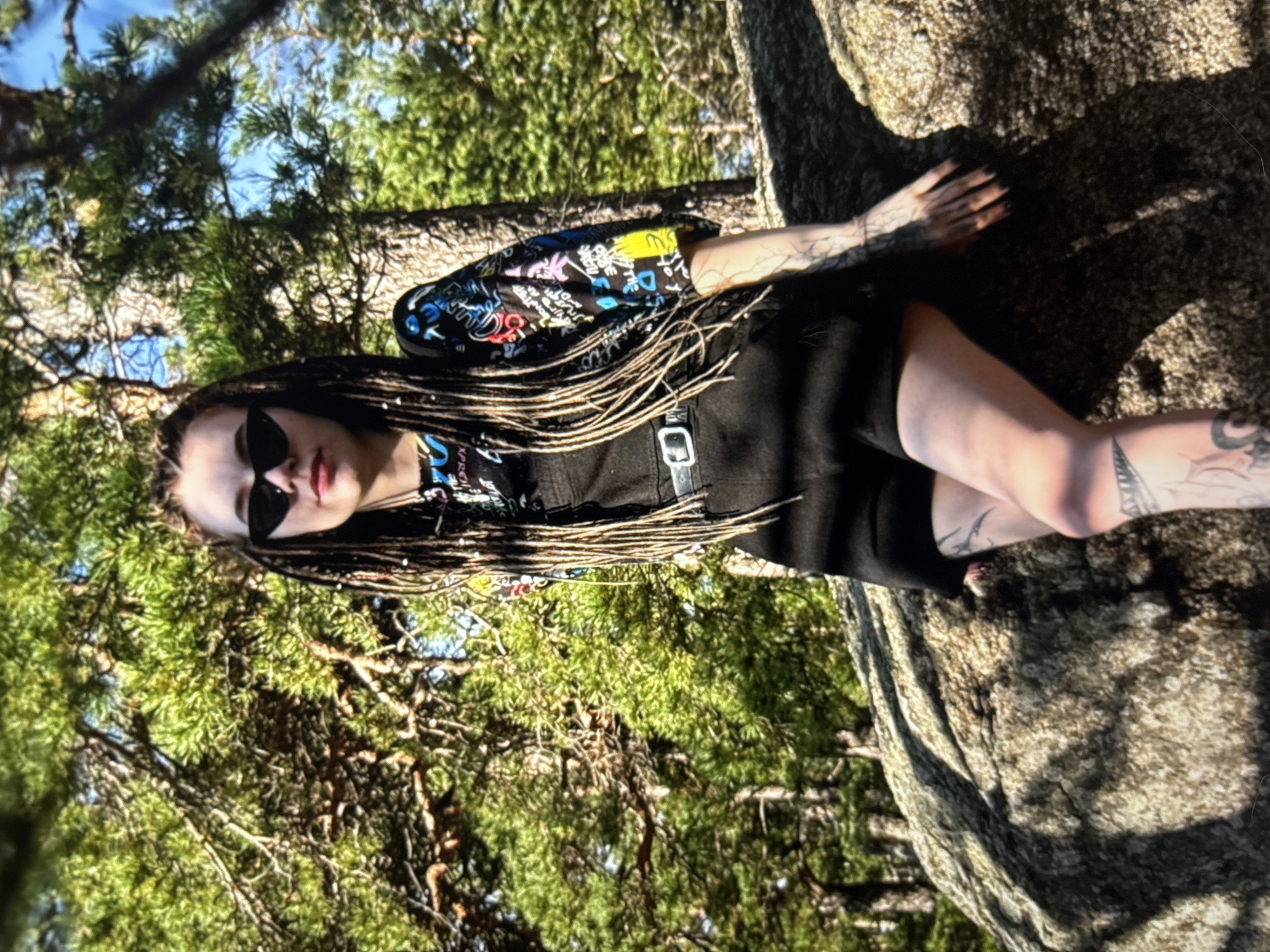 A young woman with blonde dreadlocks and cat-eye sunglasses poses leaning against a large rock in a forested area. She is wearing black overalls over a short-sleeved shirt with a colorful graffiti-style print and a black belt with a silver buckle. Her legs are visible, showing tattoos. The background consists of pine trees and a bright blue sky. The image appears to be taken in a natural outdoor setting, possibly a park or wooded area within the city of Omsk, Russia, given the context.