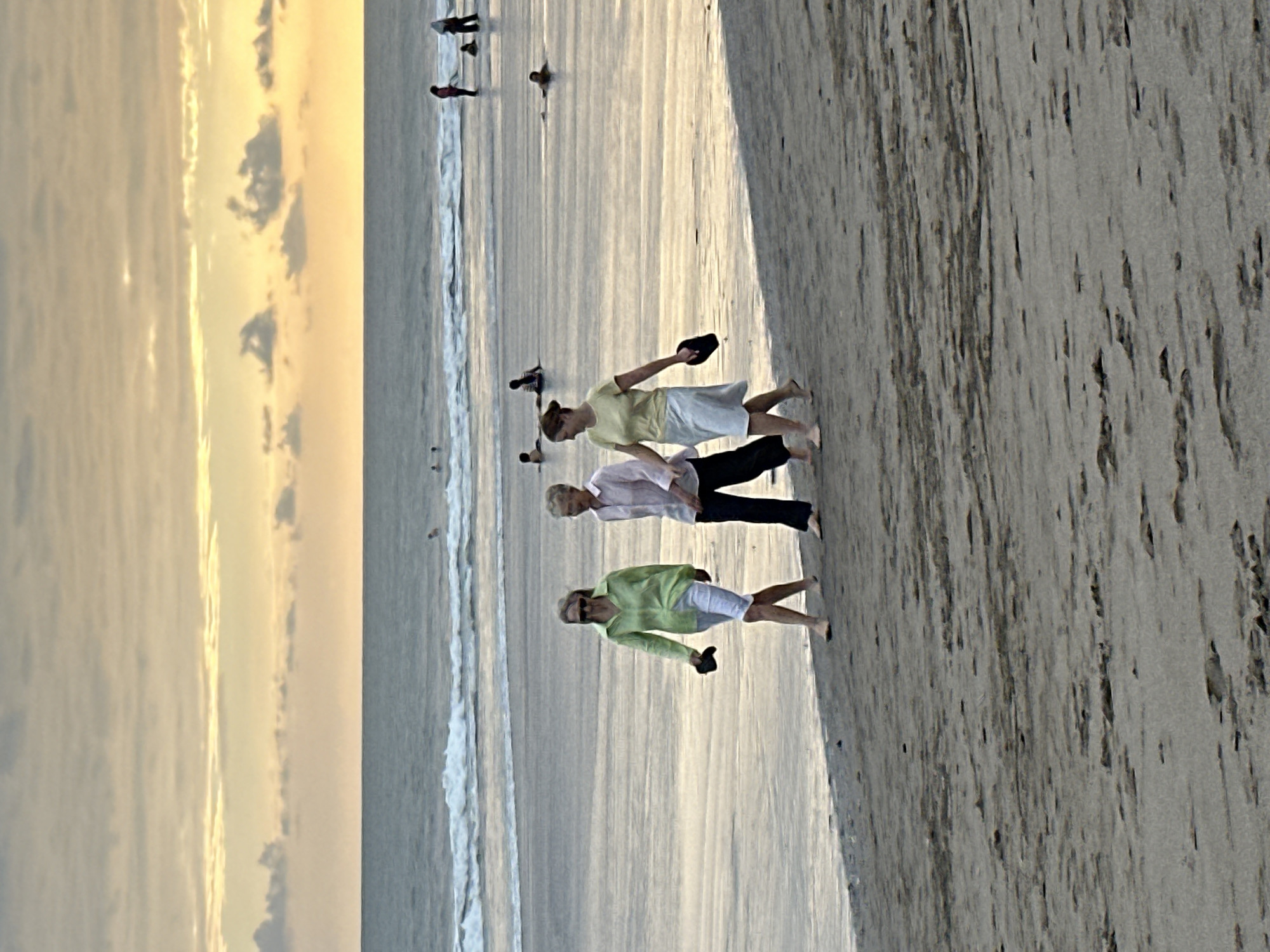 Three women are walking on a sandy beach on the coast of North Kuta, Indonesia. The woman on the left is wearing a light green shirt and white shorts, carrying a dark sandal in her right hand. The middle woman is wearing a pink shirt and dark pants, and the woman on the right is wearing a yellow shirt and a light-colored skirt, also carrying a dark sandal in her right hand. The ocean is calm with small waves, and there are several people in the water in the distance. The sky is overcast with warm yellow and orange hues near the horizon, indicating sunset. The sand is wet near the water's edge and drier further up the beach.