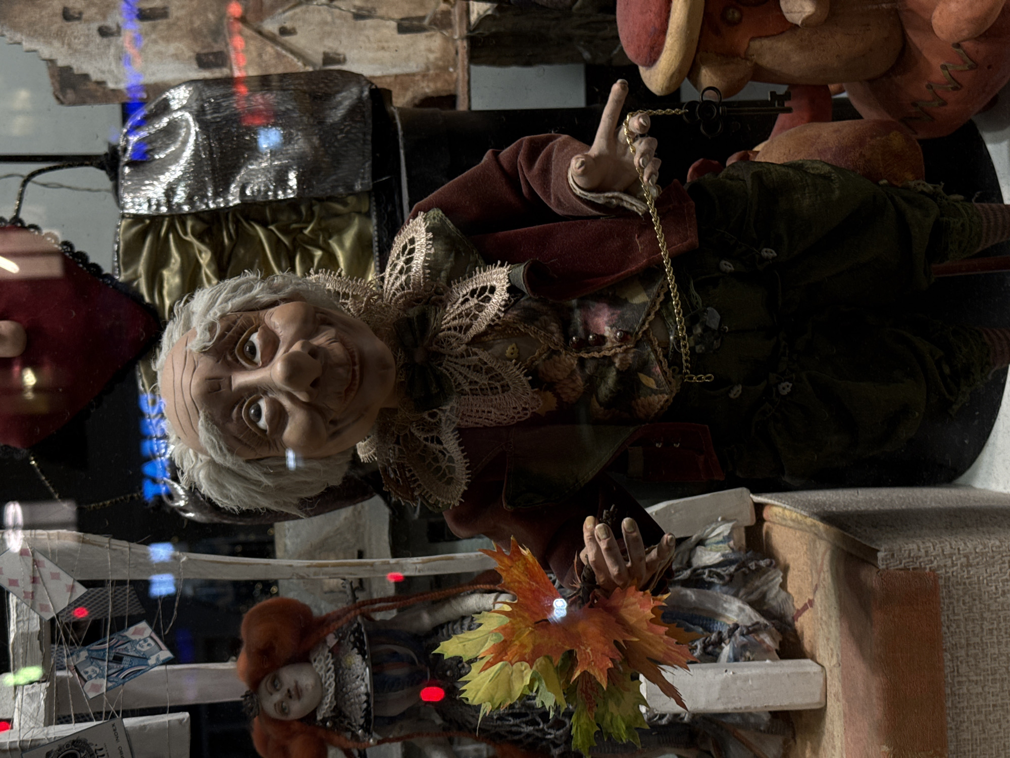In a display in the city of Moscow, Russia, a life-sized doll with pale skin and white hair is the main subject. The doll is dressed in a dark green outfit and a rust-colored velvet jacket, adorned with a lace collar. It holds a golden chain and an autumnal bouquet of leaves. Behind the doll is a silver trunk and a red hat. In front of the doll, a second doll with red hair and a lace collar is also present. Playing cards are hung up in the display, along with light cords, and the entire scene is framed by the store's shelving.