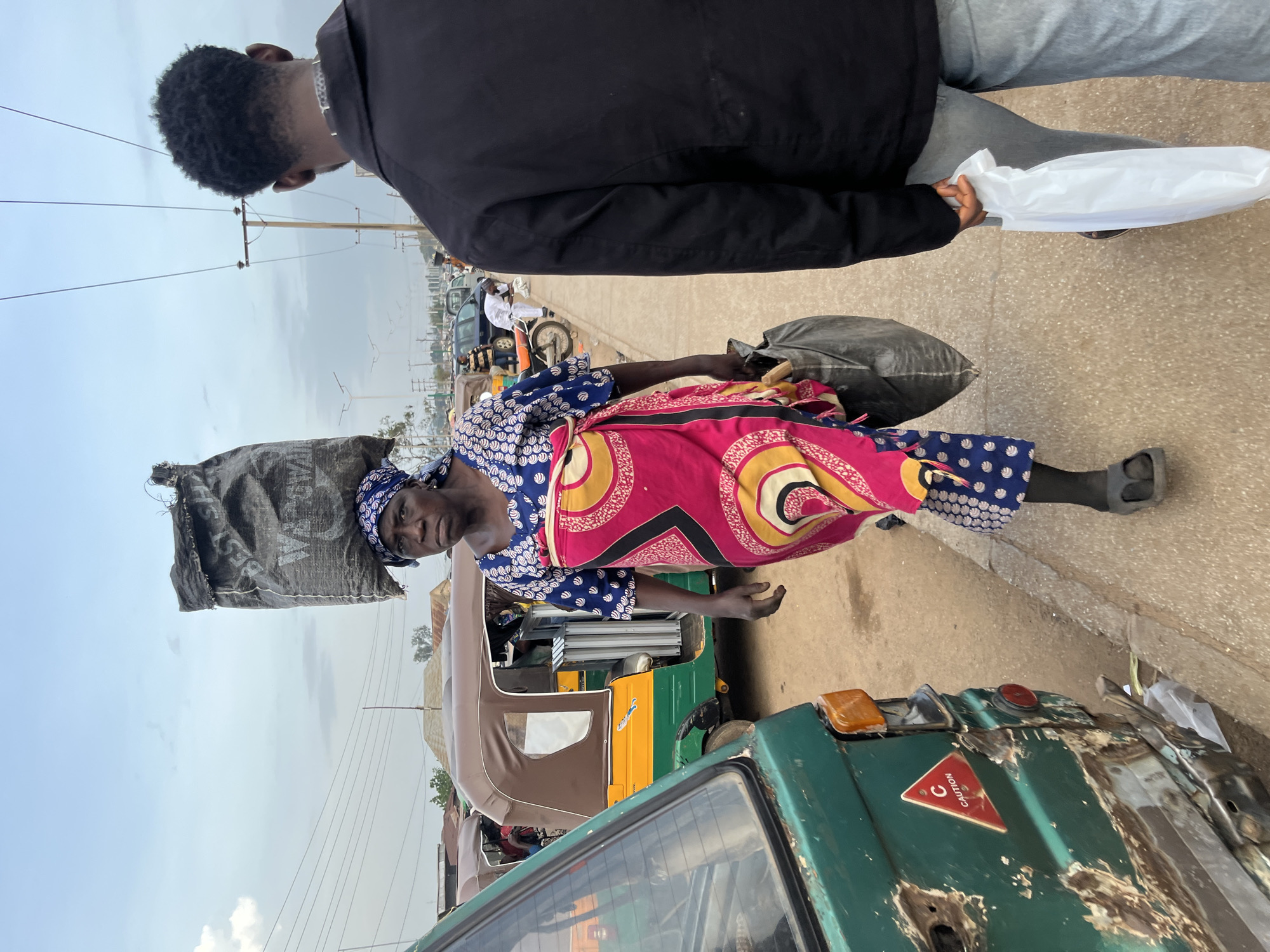 In Bukuru, Nigeria, a woman walks with a large black sack balanced on her head. She wears a blue and white patterned top and a vibrant pink and yellow wrap skirt, with dark leggings and sandals. In her right hand, she carries another black bag. The woman is walking on a dirt and gravel path beside a street lined with parked auto-rickshaws. A green vehicle with significant rust and damage is parked on the left side of the frame, displaying a red triangular "CAUTION" sticker. A man in a black jacket and jeans walks on the right side of the frame, carrying a white plastic bag. In the background, overhead wires crisscross the sky, and a variety of vehicles, including cars and motorcycles, are visible, suggesting a busy market or transportation hub. The sky is overcast with clouds.