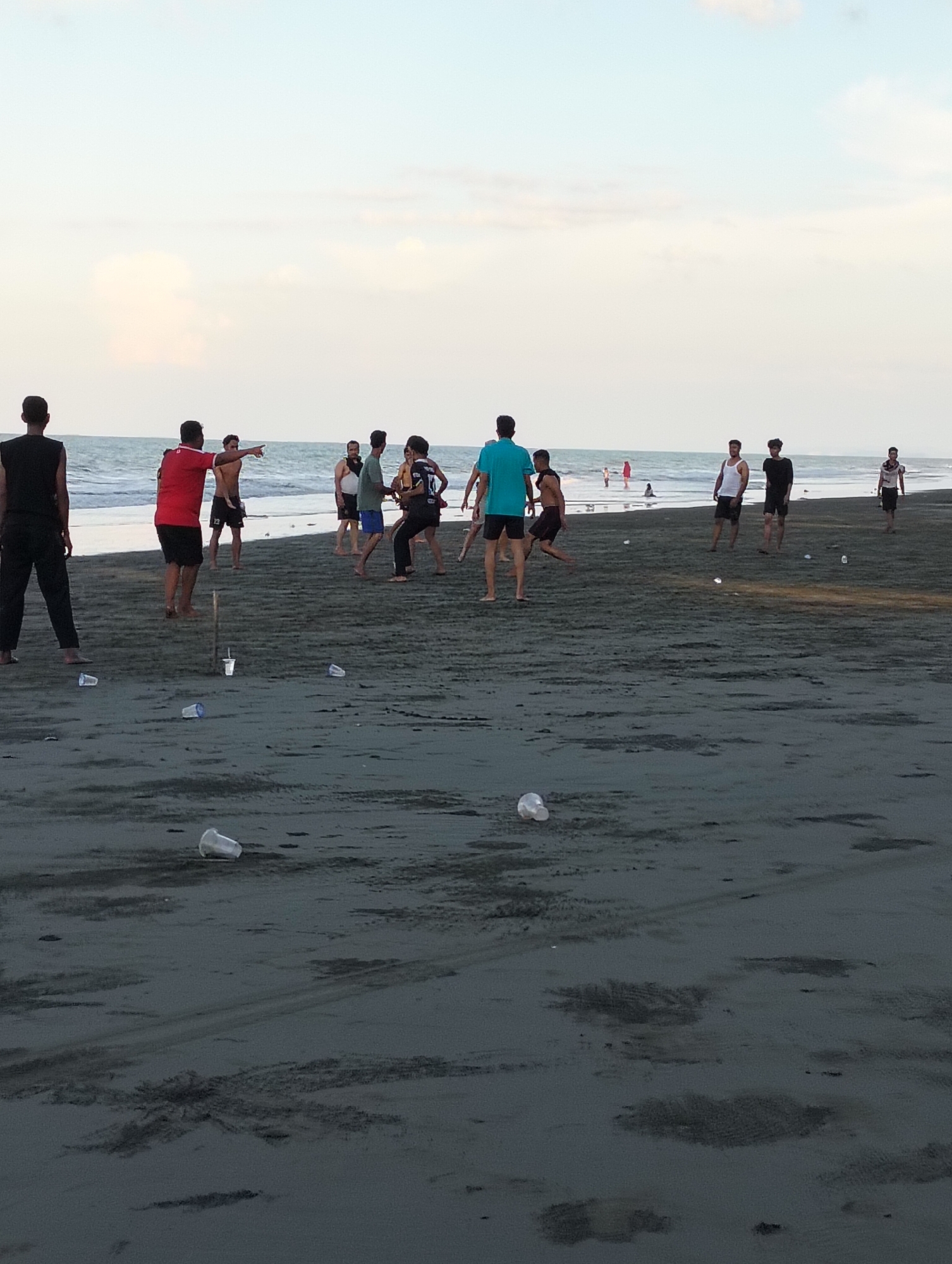 The image depicts a beach scene with several individuals on the dark sand near the water. In the foreground, disposable cups are scattered on the sand. Several people are visible playing or socializing on the beach. On the left, a person in black clothing stands facing away from the camera. Further down the beach, a person in a red shirt points towards the sea. A group of individuals are clustered in the middle ground, some appearing to be engaged in a game or activity. In the background, the ocean meets the horizon under a pale sky with scattered clouds. The overall environment is a beach with a presence of litter. This scene is consistent with the beaches found in Benteng, Indonesia, which are known for their dark sand and recreational activities.