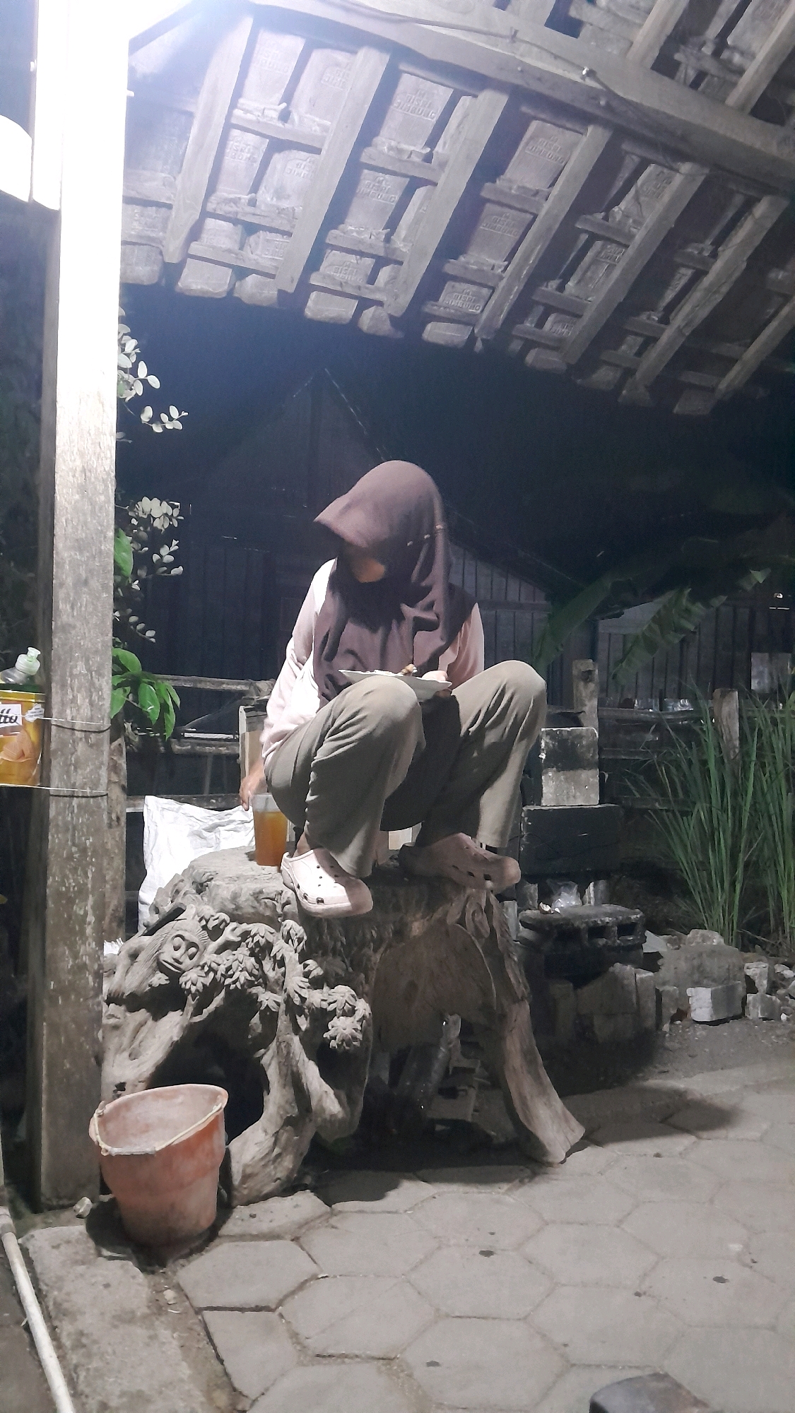 In Sumberarum, Indonesia, a person wearing a brown hijab and a light-colored shirt over green pants squats on a large, carved wooden structure. The person is holding a plate of food and a fork, and appears to be eating. A glass of amber liquid sits on the wooden structure near the person. To the left of the wooden structure, a bucket sits on the ground. The scene is outdoors, under a thatched roof supported by wooden beams. The ground is paved with hexagonal tiles. In the background, there are some plants and brick structures. The image is dimly lit, suggesting it was taken at night.