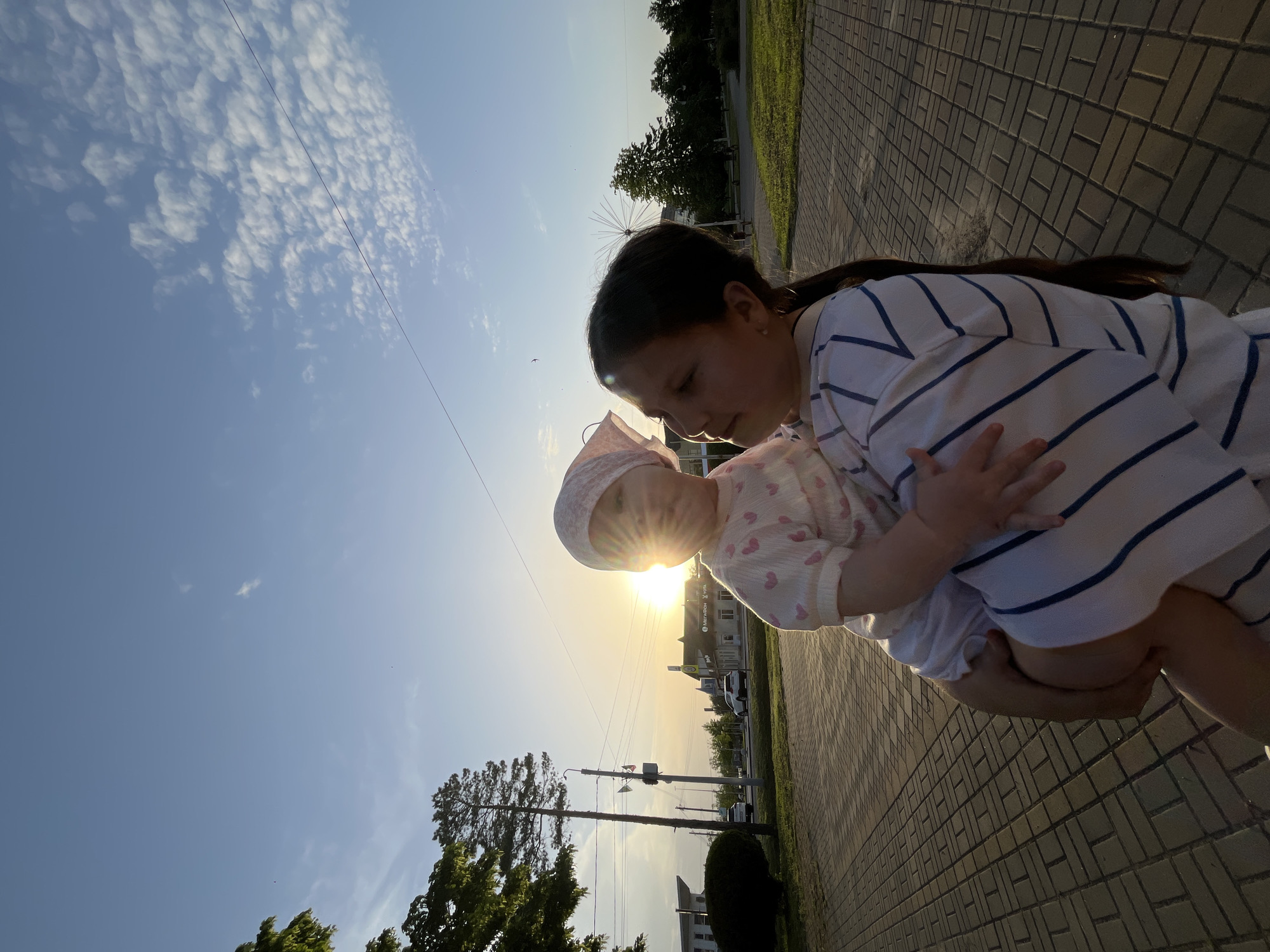 A young woman holds a baby in front of her, the setting sun behind them creating a flare effect. The woman has dark hair and is wearing a white shirt with blue stripes. The baby wears a light-colored hat and patterned clothing. They appear to be standing on a paved area. Trees and buildings are visible in the background. The scene seems to be taking place near Lenin Street and Karl Marx Street in Novopokrovskaya, Russia. Power lines and utility poles are also visible.
