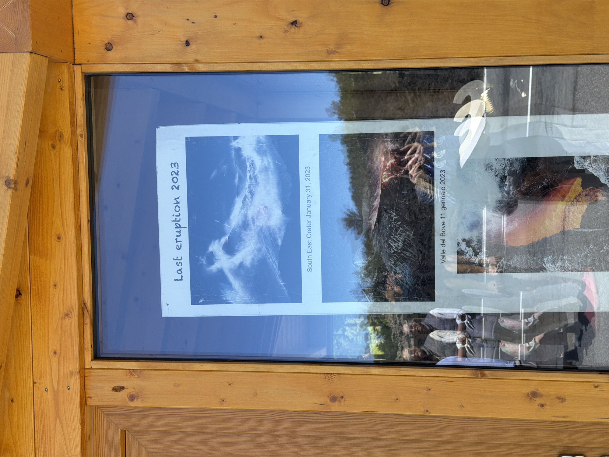 The image shows a glass-enclosed display on a wooden structure. Inside the display are two printed photographs. The top photograph depicts a sky filled with dramatic, white clouds against a blue background, with text below that reads "Last eruption 2023 South East Crater January 31, 2023". The bottom photograph shows a volcanic landscape with glowing molten rock and solidified lava, with text below that reads "Valle del Bove 11 gennaio 2023". In the reflection on the glass, two individuals are visible, wearing vests and shirts, and the photographer's hands are also reflected. The wooden structure appears to be part of a building or information kiosk, potentially in an outdoor setting given the reflections. The presence of text in Italian and the mention of "Valle del Bove" suggest the location is likely near Mount Etna, a volcano in Sicily, Italy, known for its eruptions and the Valle del Bove (Bove Valley). Linguaglossa is a town on the northern slopes of Mount Etna, making it a plausible location for such a display.