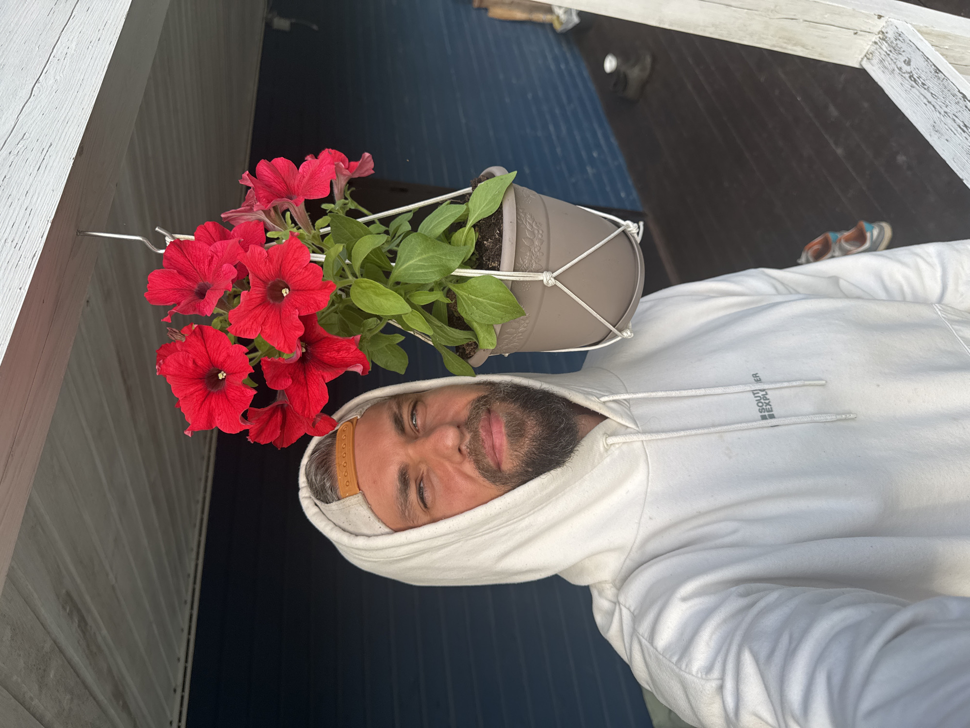 A man with a beard, wearing a white hoodie and a baseball cap, takes a selfie. To his right, a hanging pot with red petunias is suspended from the ceiling. The background consists of a dark blue wall and a wooden floor. The image is taken outdoors on a porch or balcony. The location is specified as Kuti, Russia.