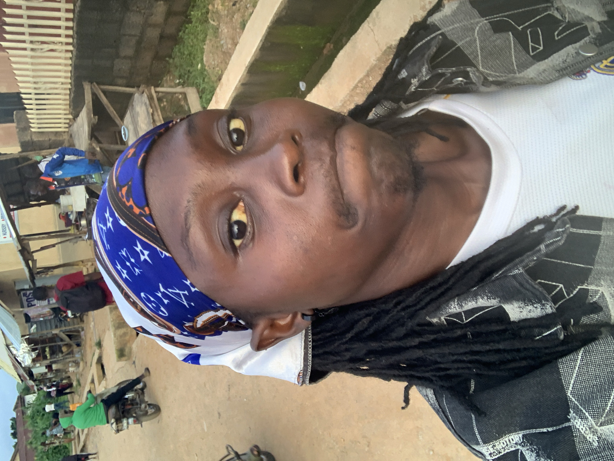 The image captures a close-up selfie of a person wearing a blue and white patterned headscarf and a black and white plaid jacket over a white t-shirt. The person has dark skin, dreadlocks, and is looking directly at the camera with a neutral expression. The background suggests an outdoor setting in Jos, Nigeria. Visible elements include a dirt road, a concrete drainage channel, a wooden structure with shelves, and buildings with corrugated metal roofs. Several other people are present in the background, some near motorbikes and stalls, indicating a bustling roadside market or street scene. A person in a green hoodie is seen riding a motorbike, and another individual in a red shirt with a backpack is walking away. The overall environment appears to be a typical urban street in Jos, characterized by its informal economy and vibrant street life.
