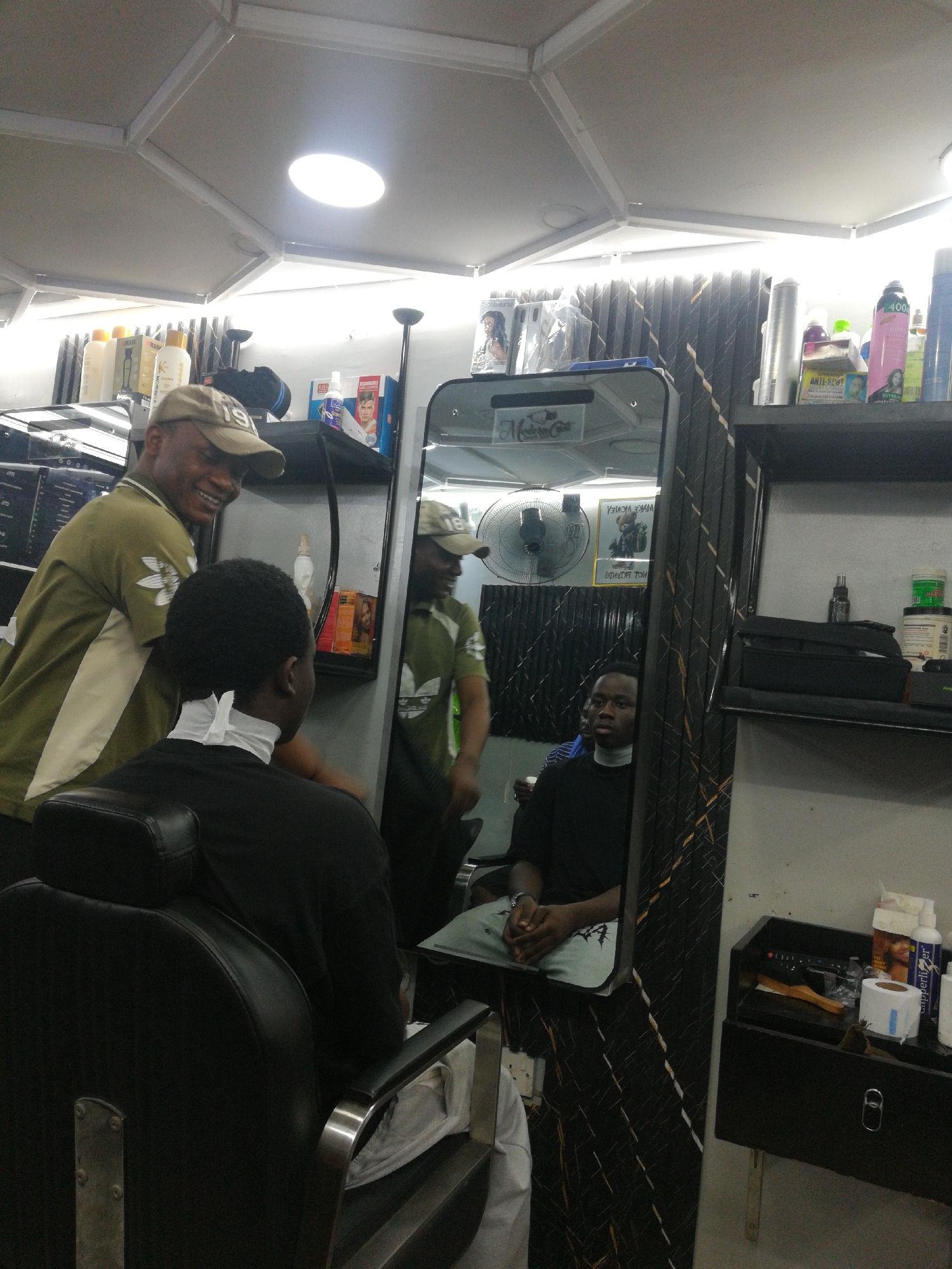 The image depicts a scene inside a barbershop. A barber, wearing a green polo shirt, is tending to a customer seated in a black barber's chair. The customer is wearing a black shirt and has a white neck strip. A mirror reflects both the barber and the customer, as well as parts of the shop's interior. The barbershop appears to be in Lagos, Nigeria, based on the context provided. The shop has shelves with grooming products, a fan, and other barbering equipment. The ceiling has a grid pattern with inset lighting.