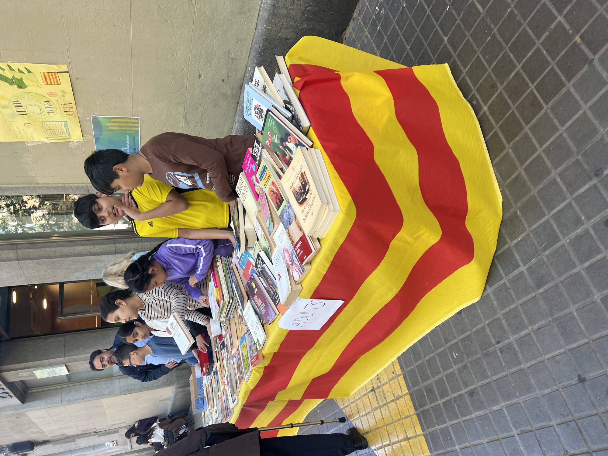 A horizontally oriented medium shot shows a book stand set up outside in Barcelona, Spain.
The book stand is covered with a flag featuring red horizontal stripes on a yellow background. It holds a selection of books. A sign reading "ADULTS" is placed near the books. Children and adults are looking at the books. Two young boys are talking next to the books, the one in the brown shirt is listening to the boy in the yellow shirt. A girl is looking at the books with a striped shirt, next to an adult, followed by two other adults. A cane is visible on the ground next to the stand. Posters are visible on the wall behind the stand.
The ground is made of concrete. The sky is visible in the background.