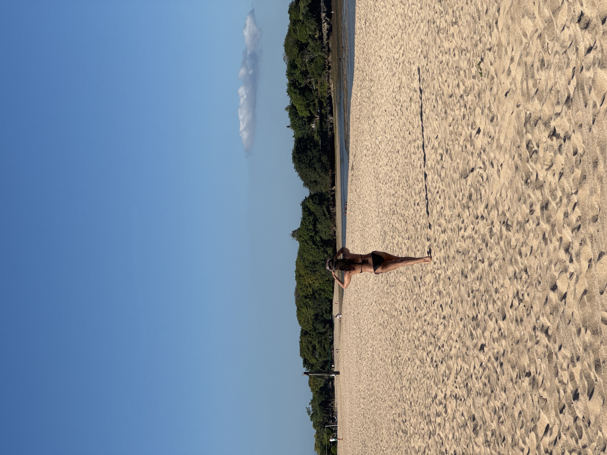The image shows a beach with a person lying on the sand. The sky is blue with a tall, thin cloud to the left. A line of trees and vegetation borders the beach. The person is lying prone and is wearing a bikini. The location is Kuta Selatan, Indonesia.