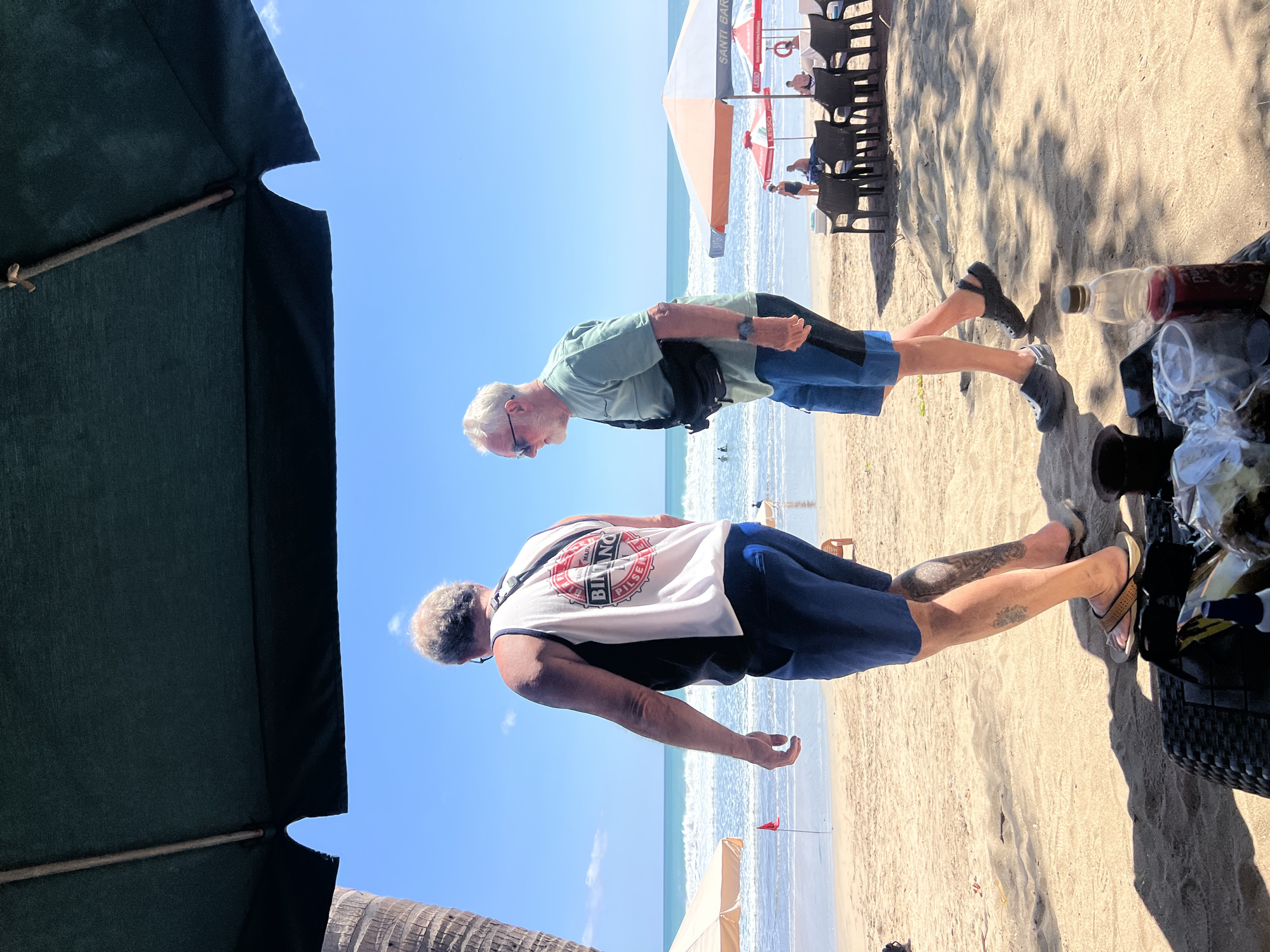 Two men are walking on a sandy beach in Kuta, Indonesia. The man on the left, facing away from the camera, wears a black and white tank top with a graphic print and blue shorts. He has tattoos on his legs and wears flip-flops. The man on the right, facing towards the ocean, wears a light green t-shirt, blue shorts, sunglasses, and dark slip-on shoes. He has a black fanny pack across his chest and a watch on his left wrist. In the background, the ocean has moderate waves, and several beach umbrellas and chairs are set up, with some people visible in the water and sitting at tables. The scene is illuminated by bright sunlight under a large, dark green umbrella in the foreground.