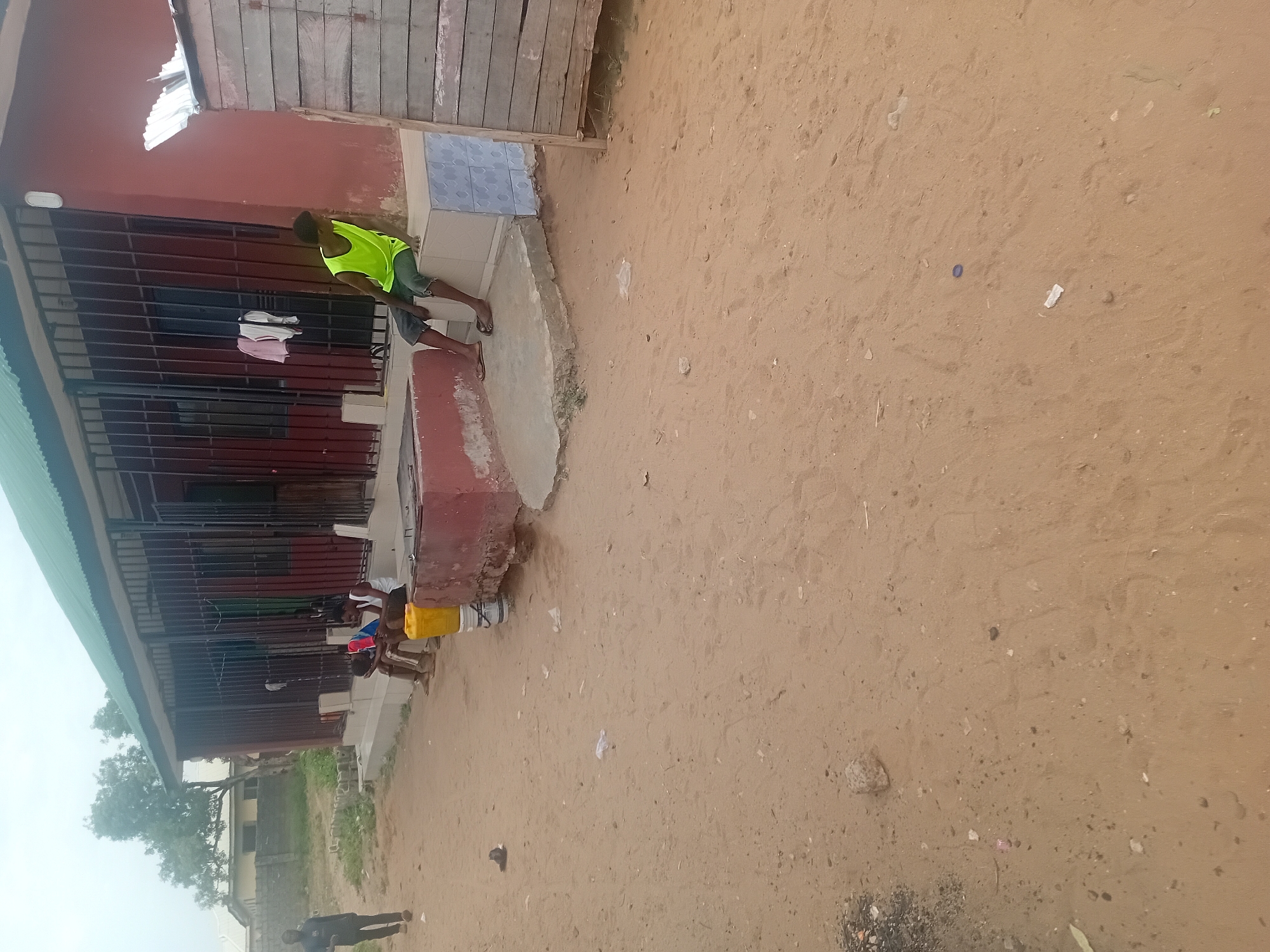 A ground-level, outdoor shot depicts a sandy expanse next to a building. The building, clad in red paint and topped with a green corrugated roof, features a barred window displaying suspended clothing items. A person wearing a neon yellow top and dark shorts stands near the building's entrance. Adjacent to the building, several individuals sit, with yellow buckets and metal containers nearby. The ground shows minimal vegetation and scattered debris. In the background, greenery and other structures are visible. The location is identified as the city of Unknown, Unknown.