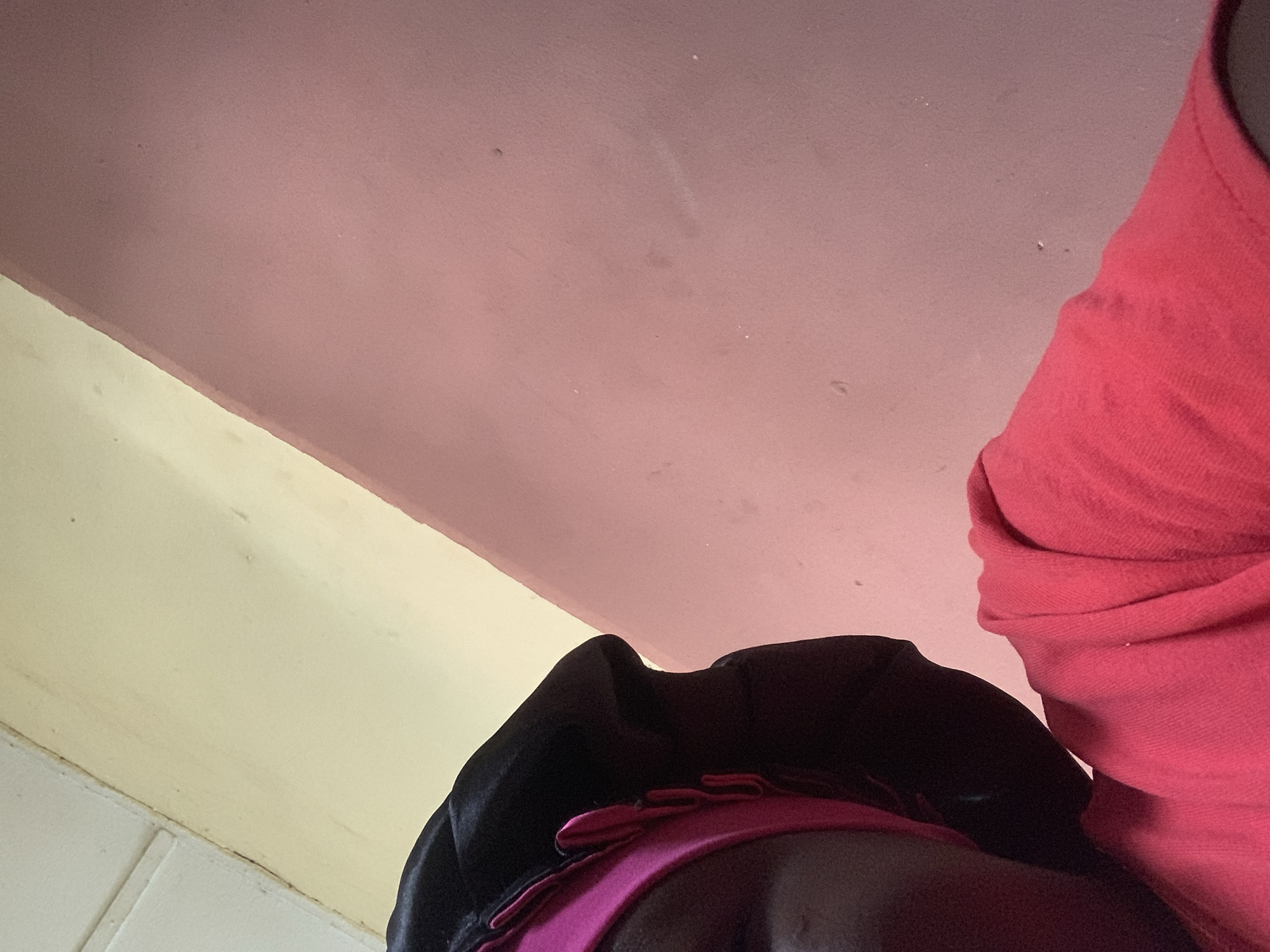 The image shows a close-up view of a person wearing a black satin bonnet with a pink trim and a red t-shirt. The person's face is partially visible on the left side of the frame. The background consists of a wall painted in two different colors: a light yellow or cream color at the top and a pinkish-red color below. The lighting suggests an indoor setting, possibly a home. The scene is static and focuses on the individual and their immediate surroundings. The specific location in Jauro Yinu, Nigeria is not discernible from the visual information presented.