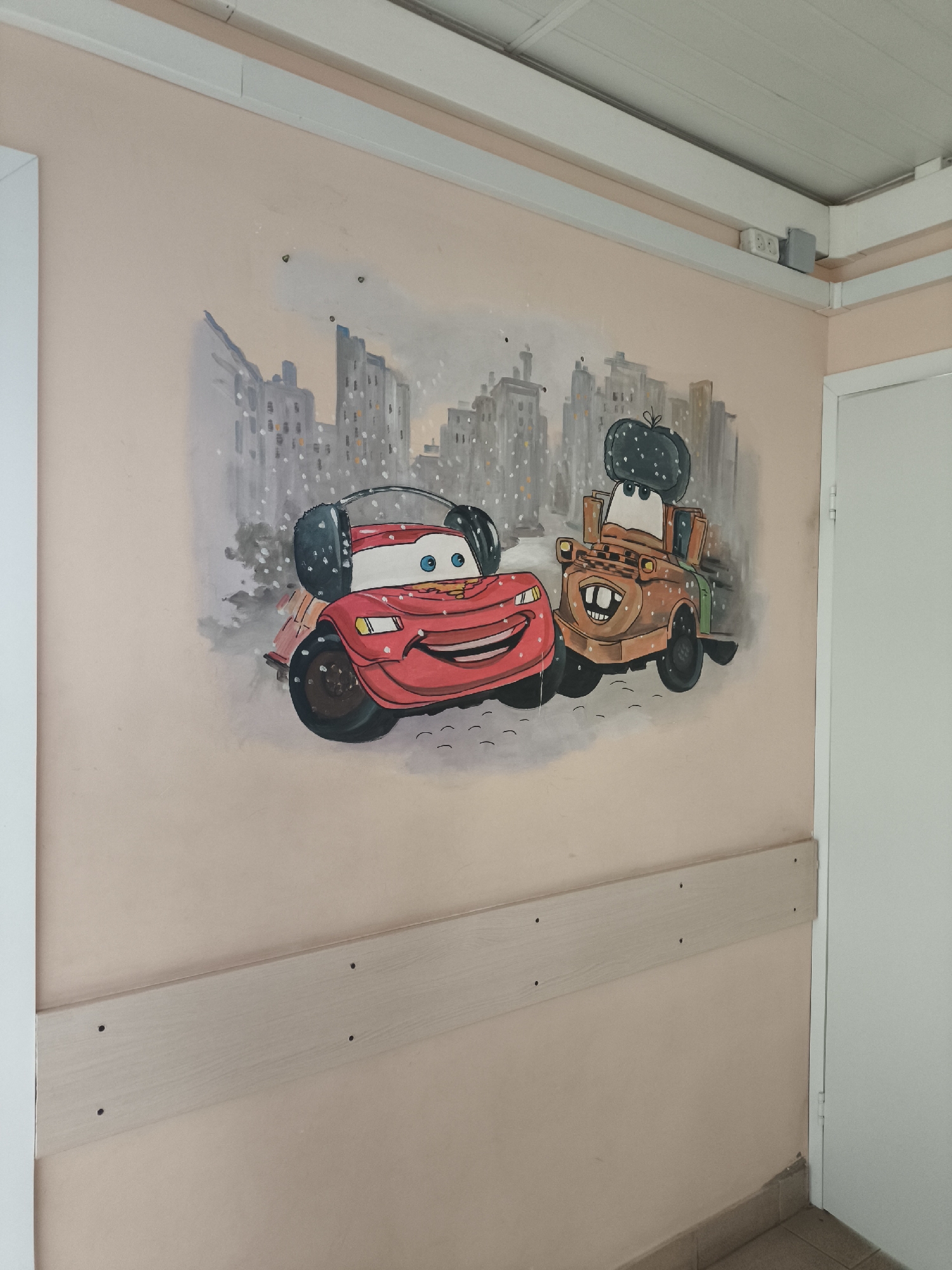 A wall mural of cartoon cars is painted on a peach-colored wall. The left side of the mural shows a red car with headphones and a white roof. The right side of the mural shows a brown tow truck wearing a dark fuzzy hat. Behind the two cars is a city scene with tall buildings and what looks like snow. Below the mural is a light tan bumper rail. The mural is in a room with white trim and a white door to the right. The location is not discernible from the image.