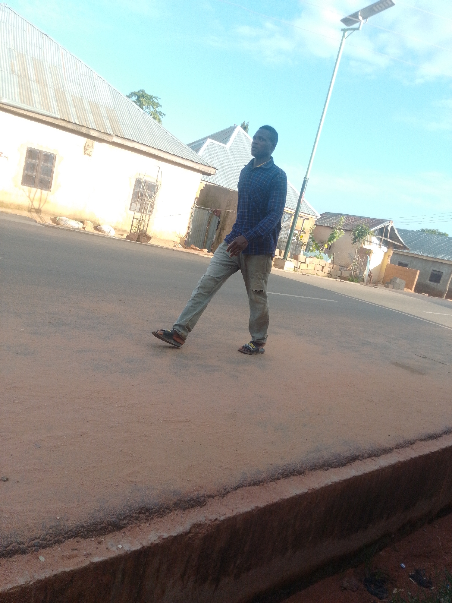 A young African man is standing outdoors in front of a row of buildings. He is wearing a blue and black checkered shirt and light-colored pants. He is looking to the left and his body is angled to the right. A lamppost with a solar panel is visible behind him. The background consists of buildings with corrugated metal roofs and a clear blue sky. The ground in the foreground is sandy and reddish-brown. This scene is consistent with an outdoor setting in the city of Gombe, Nigeria.