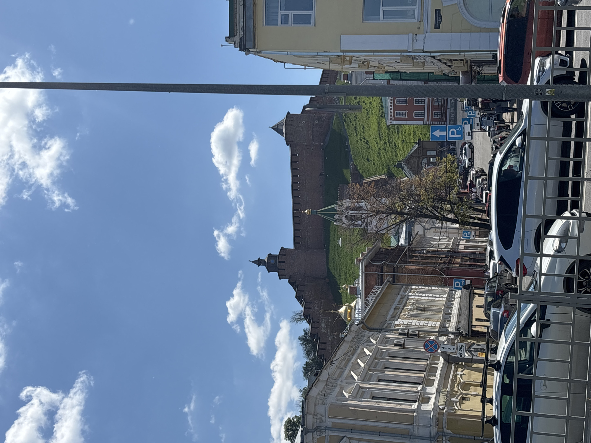 The image presents a daytime urban landscape in Nizhny Novgorod, Russia, showcasing historical architecture alongside a modern street scene. The composition is effectively in landscape orientation when viewed correctly.
In the foreground, a street is lined with parked cars, including a white SUV/crossover, a dark car, and a red car, partially obscured by a metal fence or railing on the right. To the left of the street are multi-story buildings displaying classical architectural elements with ornate windows and decorative facades. A tall, dark lamppost or utility pole runs vertically through the middle ground, partially dividing the scene.
The midground is dominated by a majestic, red-brick Kremlin wall, complete with watchtowers, rising from a steep, grassy hillside. One prominent tower features a distinctive pointed roof. Below the Kremlin wall, nestled among trees, a building with a gleaming golden dome is visible, likely a church or cathedral.
The background is a clear, bright blue sky dotted with scattered white cumulus clouds. The strong sunlight and visible shadows indicate a bright, sunny day, likely mid-morning or early afternoon. The scene is devoid of people, contributing to a sense of tranquility.
Visible text includes several blue parking signs with a white "P" and arrows pointing in various directions. There is also a red circular "No Stopping" sign with a blue cross and an arrow, along with other symbols, near the street-level buildings. The overall scene captures a picturesque blend of historic Russian fortifications and contemporary city life.