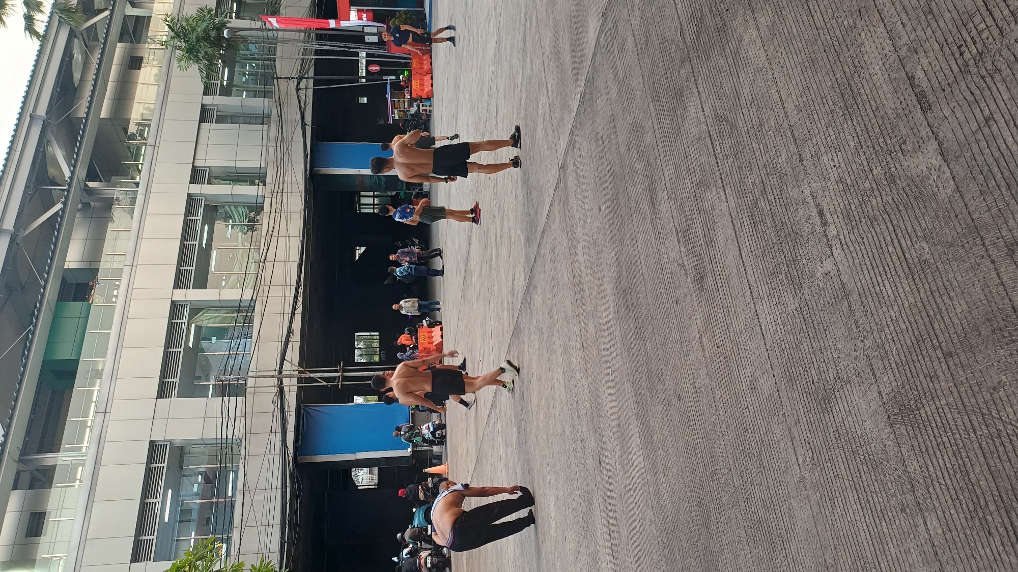 The image shows an outdoor recreational area or parking lot with a modern building in the background. Several men are visible in the foreground and midground, some of whom are shirtless and wearing athletic shorts. One man in the lower left corner is stretching, bent over with his hand on his shoe. Other men appear to be standing or walking. In the midground, there are several parked motorcycles and a few individuals in the background observing. The building behind them has multiple levels with windows and balconies. The ground is paved with a textured concrete surface. The scene suggests a casual, possibly athletic, gathering in a semi-urban environment. The presence of a red and white Indonesian flag on a pole indicates the location is in Indonesia. This scene is likely situated in an area accessible to the public, possibly near a sports facility or community center within South Jakarta, Indonesia.