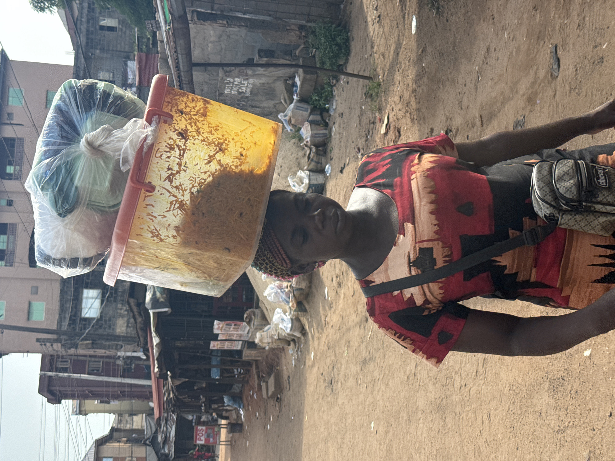 A woman walks along a sandy road in Onitsha, Nigeria. She balances a large, orange plastic container filled with cooked rice, topped with a bag of unknown items covered in plastic wrap, on her head. She wears a patterned dress in shades of red, brown, and black, and a small bag is slung across her body. The background shows a bustling urban scene with buildings, open-air businesses, and accumulated trash along the roadside.