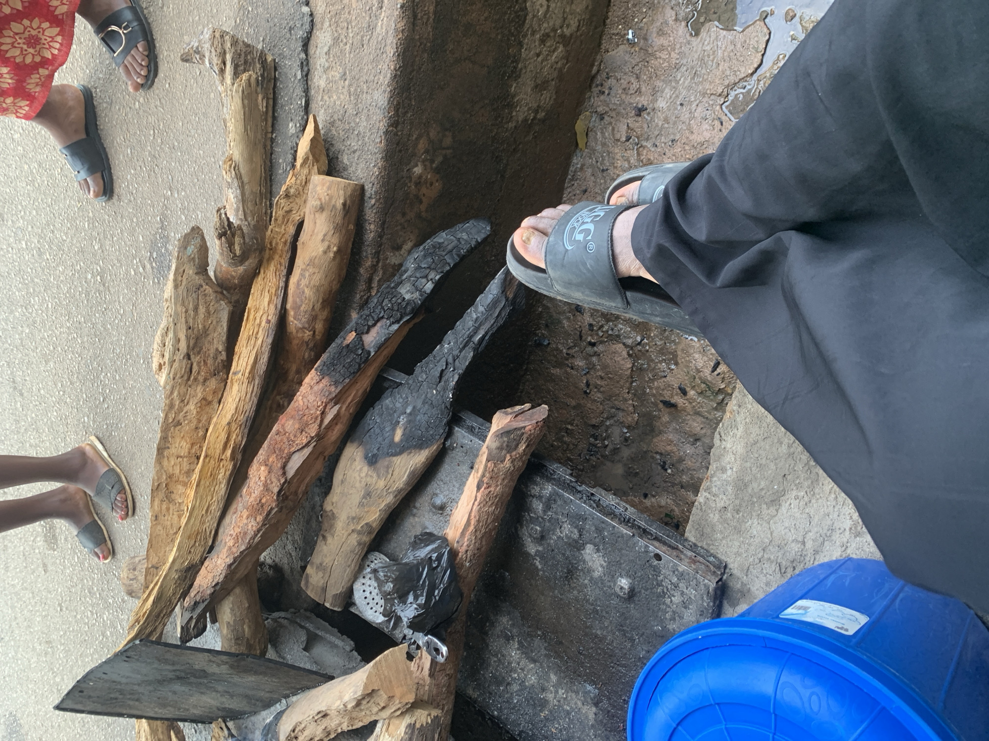 The image depicts an outdoor scene likely in the city of Jos, Nigeria, focusing on a pile of firewood. In the foreground, a person's legs clad in black trousers are visible, wearing grey slides. The firewood, a mix of burnt and unburnt logs, is arranged on a dark, wet surface, possibly a metal grate or tray. To the left, a bright blue plastic container is partially visible. In the background, the lower legs and feet of two individuals are seen standing on a paved surface. One person wears grey slides, and the other wears black slides with a gold accent. The overall environment appears to be a somewhat gritty, outdoor location.