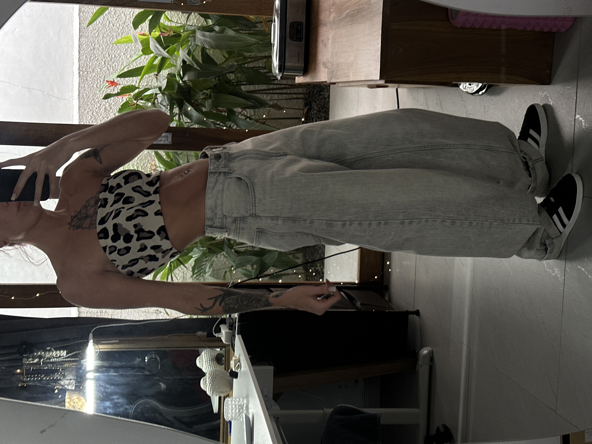 A woman stands indoors, wearing a leopard-print top and baggy jeans with black and white sneakers. She is holding a black smartphone to her ear. The background includes a mirror, a vanity, and indoor plants near a window. The location is in Dalung, Indonesia.