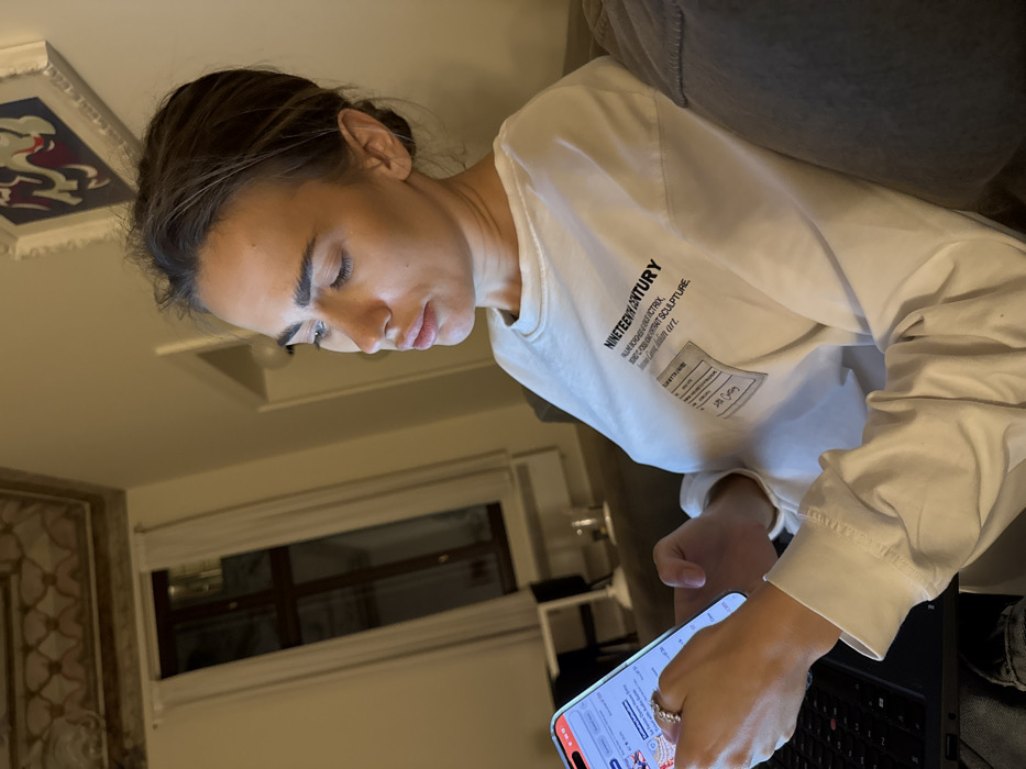 An indoor scene in Venice, Italy, depicts a female subject seated, illuminated by the screen of a smartphone held in her left hand. She has dark hair styled away from her face and wears a white long-sleeve shirt with "NINETEENTH CENTURY" and other text printed on the chest. A metallic ring with multiple stones is visible on her left ring finger. A black laptop is positioned in the lower left foreground. The background features light-colored walls and a tall window with dark framing and sheer white curtains, reflecting interior light. Above, the ceiling displays an elaborate, painted or frescoed pattern in geometric and organic motifs, characteristic of historical Italian architecture. A framed artwork hangs on the wall in the upper right quadrant.