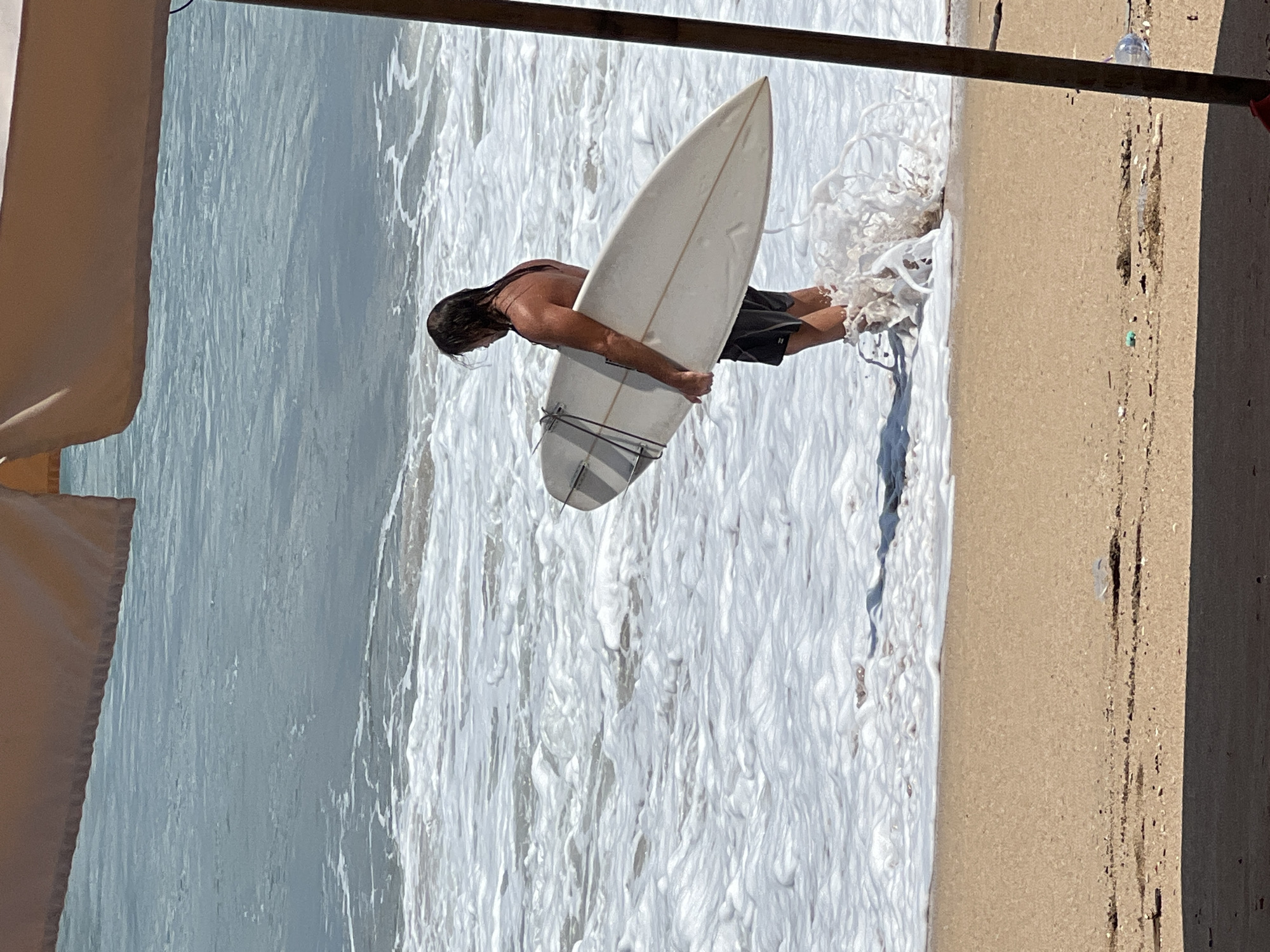 A surfer is standing in the shallow water at Kuta Beach, Indonesia, holding a white surfboard. The surfer, a male with long, wet hair, is wearing dark swim trunks. He is facing away from the camera, towards the ocean. The water is foamy and there are small waves. The beach is sandy, and there is a shadow in the foreground, suggesting the presence of a large umbrella or awning, partially visible in the top and right edges of the frame. Some small debris is scattered on the sand.