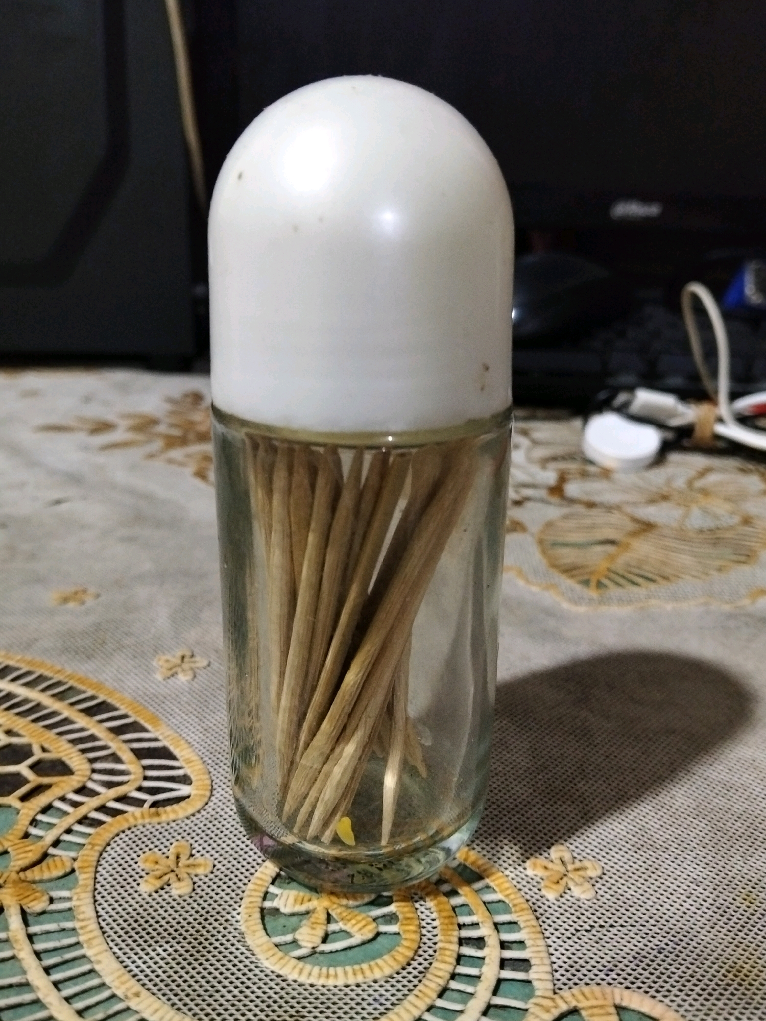 A glass jar filled with wooden toothpicks and topped with a white rounded cap sits on a patterned tablecloth. The jar is transparent, allowing the toothpicks to be visible inside. The background is dark and out of focus, with some electronic devices and cables present. The tablecloth has a woven texture with a floral design in shades of yellow and cream. The image appears to be a close-up, still life shot, taken indoors. There is no indication of the city of Teluk Pambang, Indonesia.