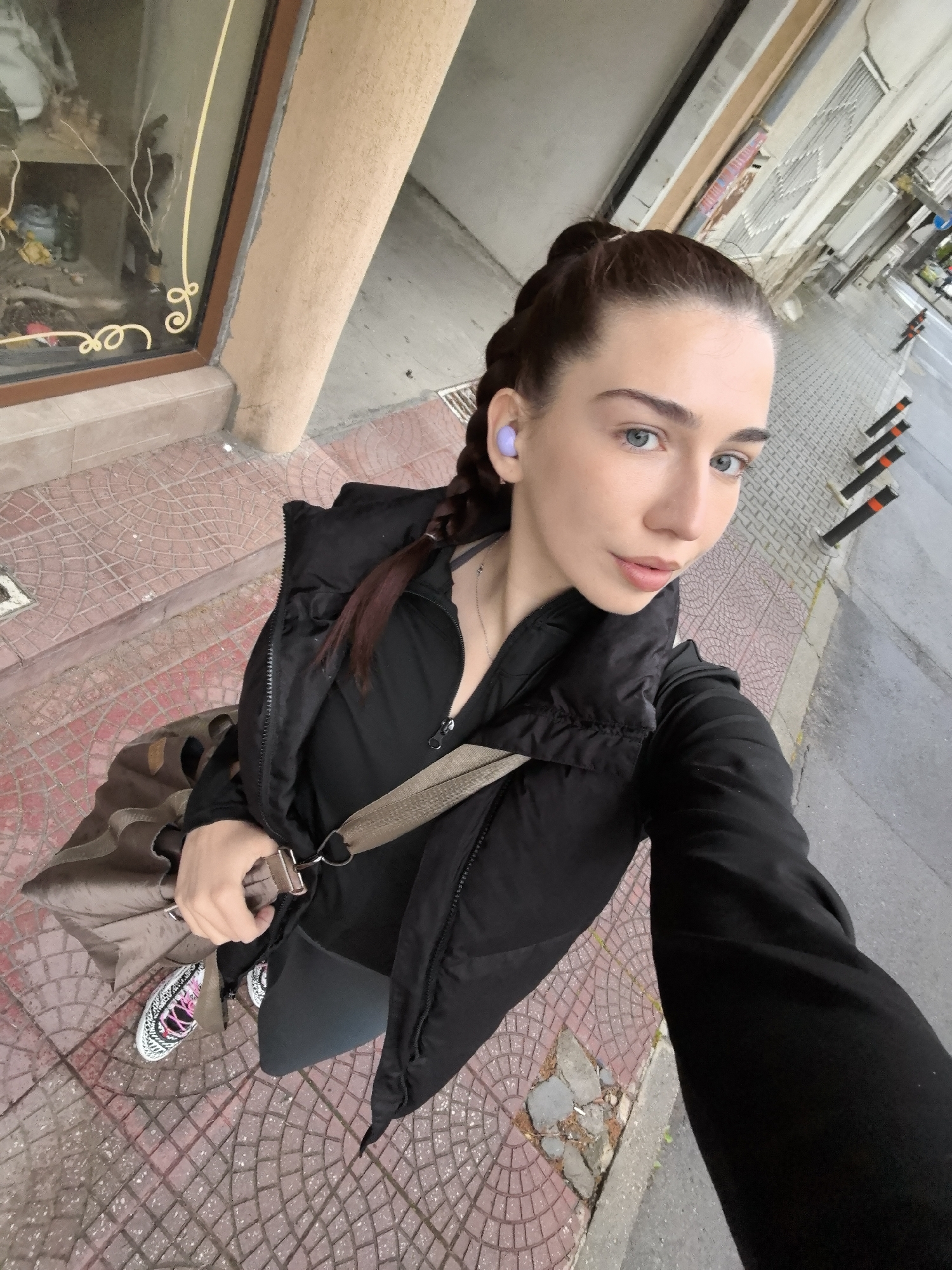 A young woman takes a high-angle selfie on a tiled sidewalk in an urban setting, likely Stara Zagora, Bulgaria. She has light skin, blue eyes, and dark brown hair styled in braids pulled back from her face. She wears a black long-sleeved top, a black puffer vest, dark leggings, and white sneakers with black patterns and pink accents. A light purple earbud is visible in her left ear, and she wears a thin silver necklace. She carries a brown/khaki duffel bag slung over her left shoulder, with her right hand resting on the strap.
The time of day appears to be daylight. To her left, there is a building with a storefront window displaying various items, framed by a brown border. The window is adorned with decorative golden-yellow lines that form stylized loops and cursive-like shapes. The adjacent building wall is light brown. The sidewalk where she stands is made of reddish-brown tiles with a patterned surface.
To her right, a curb separates the sidewalk from an asphalt road. In the background, across the street, more buildings are visible, along with several black and orange bollards or traffic cones lining the roadside. The overall scene depicts a casual moment in a city street environment.
