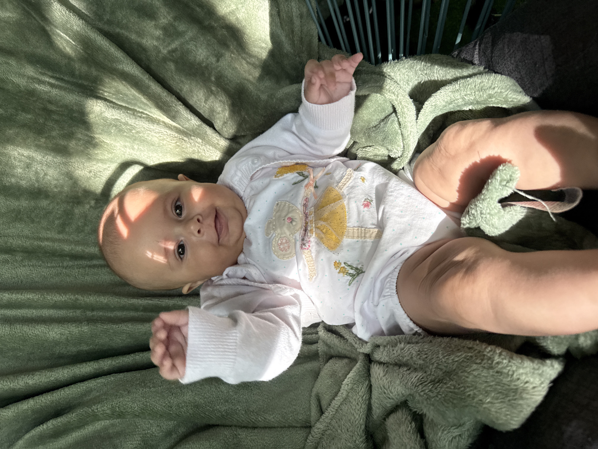 A baby is lying on a green blanket. The baby is wearing a white outfit with a pattern of flowers and a yellow dress. The baby has their right arm raised. Sunlight is casting shadows on the baby and the blanket. The background is a green blanket that is draped. There is a wire fence visible on the right side of the image. The image is taken outdoors. The location is not specified in Plovdiv, Bulgaria.