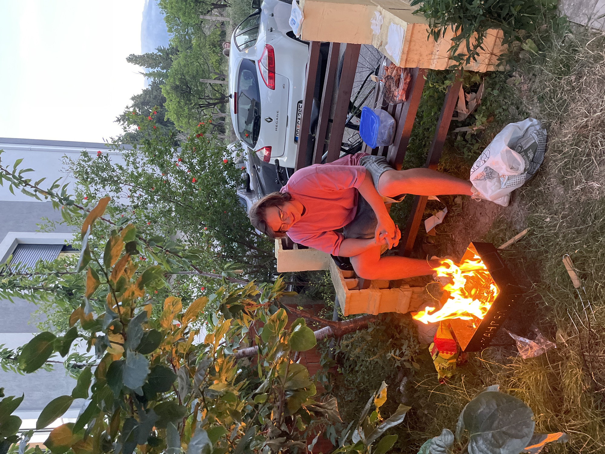 This image depicts an outdoor evening scene, likely in a backyard or garden in Bar, Montenegro. The primary subject is a person, appearing to be a young adult with glasses, wearing a pink long-sleeved shirt and shorts. They are seated on a wooden bench, looking towards the viewer with a relaxed expression.
In the mid-ground, a metal fire pit emits bright flames, indicating a bonfire or barbecue activity. A grill with food skewers rests on the bench next to the person, accompanied by a blue plastic container. On the ground near the fire pit, a white plastic bag and a bag possibly labeled "KOHLE" (charcoal) are visible. The bench itself seems to be partly constructed from or adjacent to light-colored building materials or wooden pallets.
The background features a white Renault Megane car with the license plate "DR CA 2CE", and another vehicle partially obscured. A modern white building with horizontal blinds is visible behind the car. The scene is framed by various green trees and bushes, some with small red blossoms (possibly pomegranate). Distant mountains are silhouetted against a pale, twilight sky, confirming the evening time. Another person's face is subtly visible in the far background near the car. The overall impression is one of a casual, comfortable outdoor gathering.
