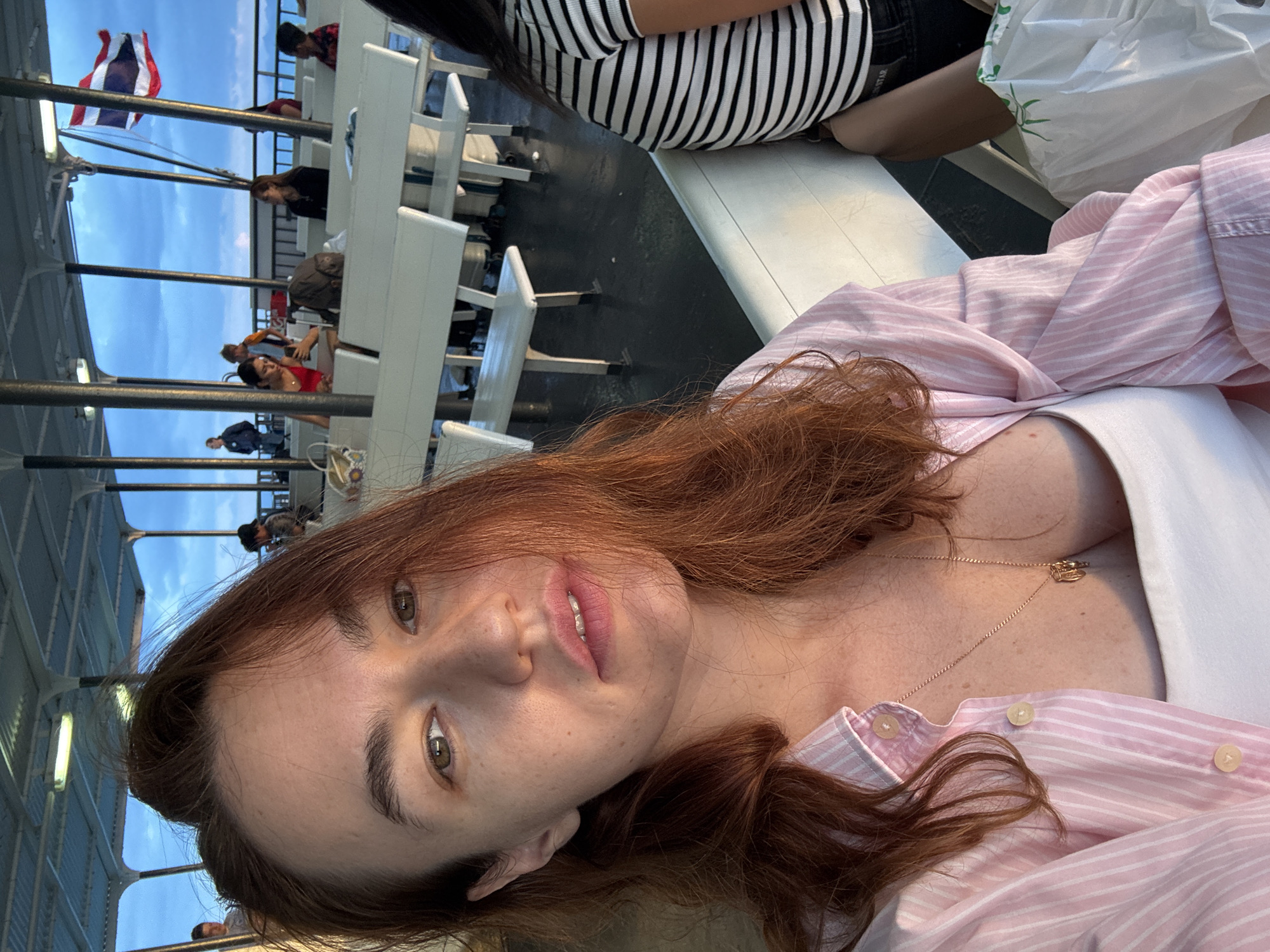 A young woman with long, reddish-brown hair is taking a selfie on a ferry. She is wearing a pink and white striped open button-down shirt over a white tank top and a gold necklace with a pendant. Her hair is partially obscuring her face, and she is looking at the camera with her mouth slightly open. In the background, other passengers are seated on white benches on the open-air deck of the ferry. The sky is blue with some clouds, and a Thai flag is visible on a flagpole. The deck appears wet, possibly from sea spray.