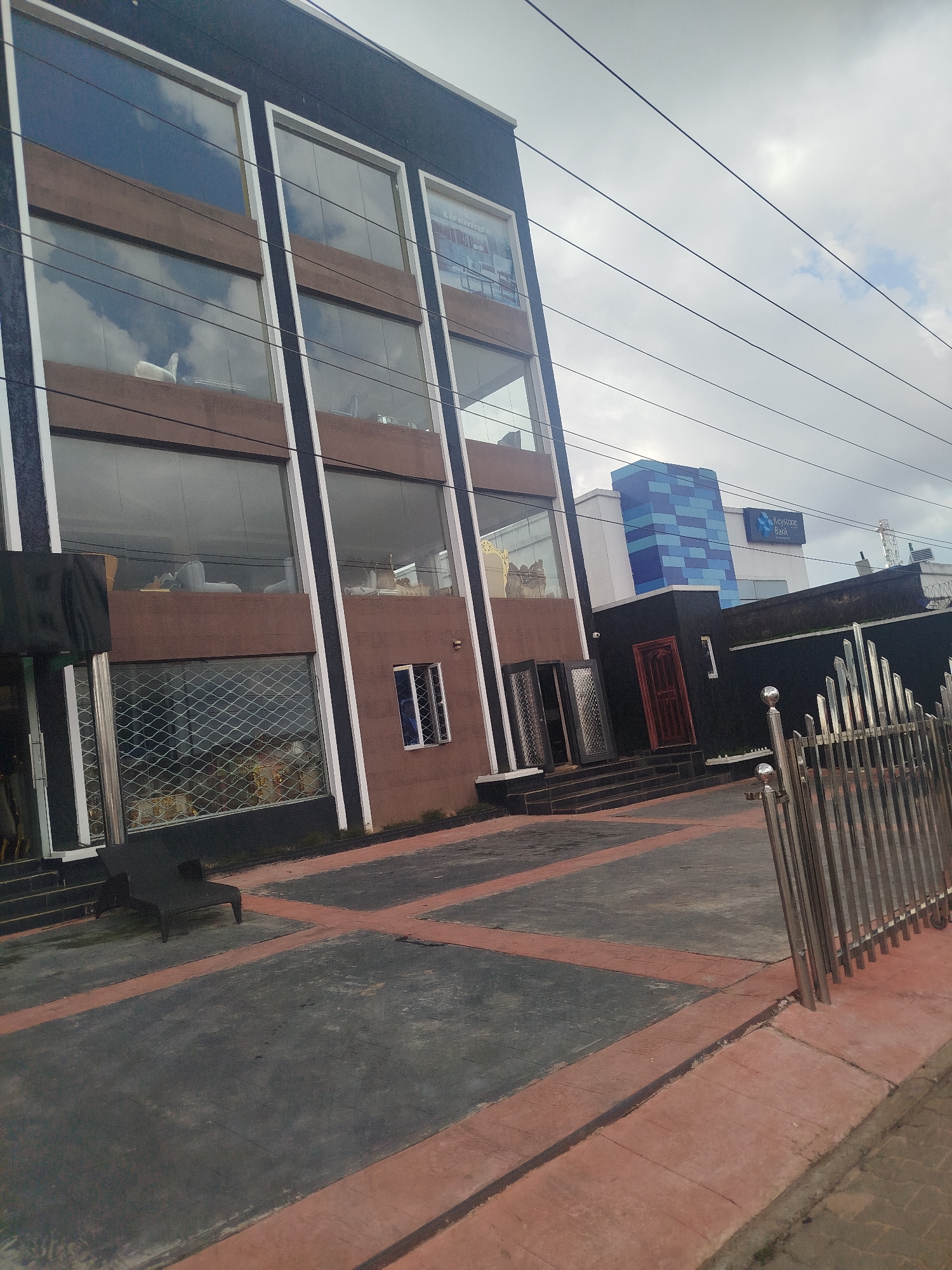 The image shows the exterior of a building in Benin City, Nigeria. The building has a dark facade with multiple levels of large windows. The lower level of the building features a shuttered storefront with a metal grate. Adjacent to this, there are double doors leading into the building, with steps leading up to them. Further to the right, a red door is visible. Behind the main building, a structure with blue and white rectangular patterns can be seen, with a sign that reads "Keystone Bank". A metal fence with vertical bars is present in the foreground on the right side of the image. The ground in front of the building is paved with dark grey squares outlined in red. A black bench-like object is positioned on the paved area to the left. The sky is overcast with some clouds.