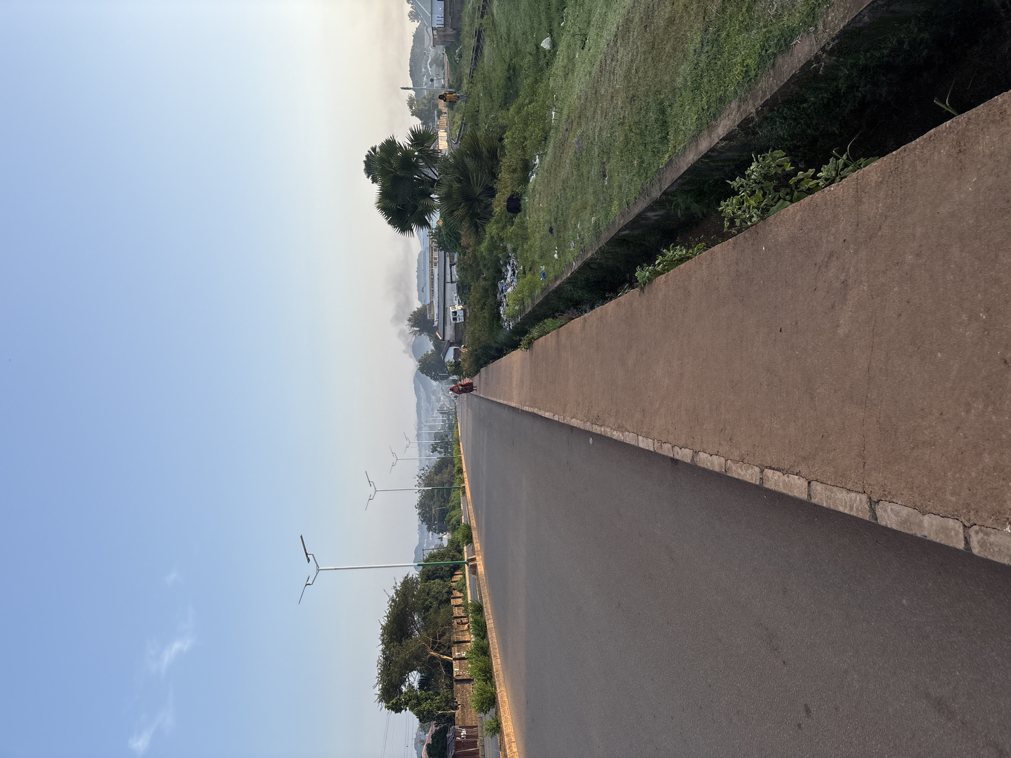 A wide asphalt road with a concrete sidewalk runs diagonally through the frame from the lower left to the upper right, suggesting it is a main thoroughfare within the city of Bukuru, Nigeria. The sidewalk is bordered by a concrete drainage ditch on the right. A few streetlights are visible along the side of the road. Beyond the sidewalk and ditch, there is a grassy area with scattered debris, including plastic waste. Further in the background, there are several trees, buildings, and distant hills. Two individuals are walking on the sidewalk in the distance, and one person is visible on the grassy area. The sky is clear with a few wisps of clouds, and the lighting suggests it is early morning or late afternoon.