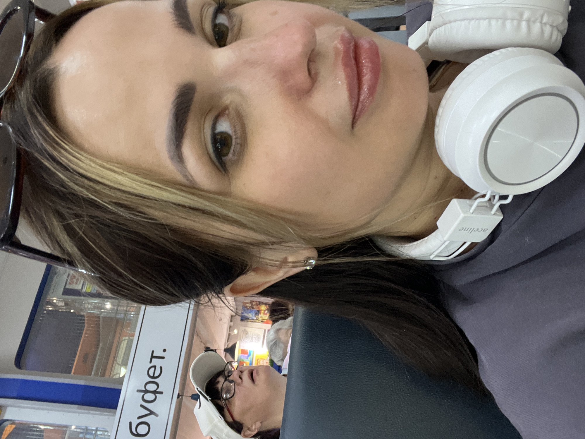A woman wearing headphones around her neck takes a selfie in what appears to be a public area, possibly an airport or train station, with a sign reading "БУФЕТ" (buffet) in the background. In the background, another woman wearing a white baseball cap and glasses looks upwards. The main subject's face is the primary focus, with her eyes looking towards the camera. The lighting suggests an indoor environment.