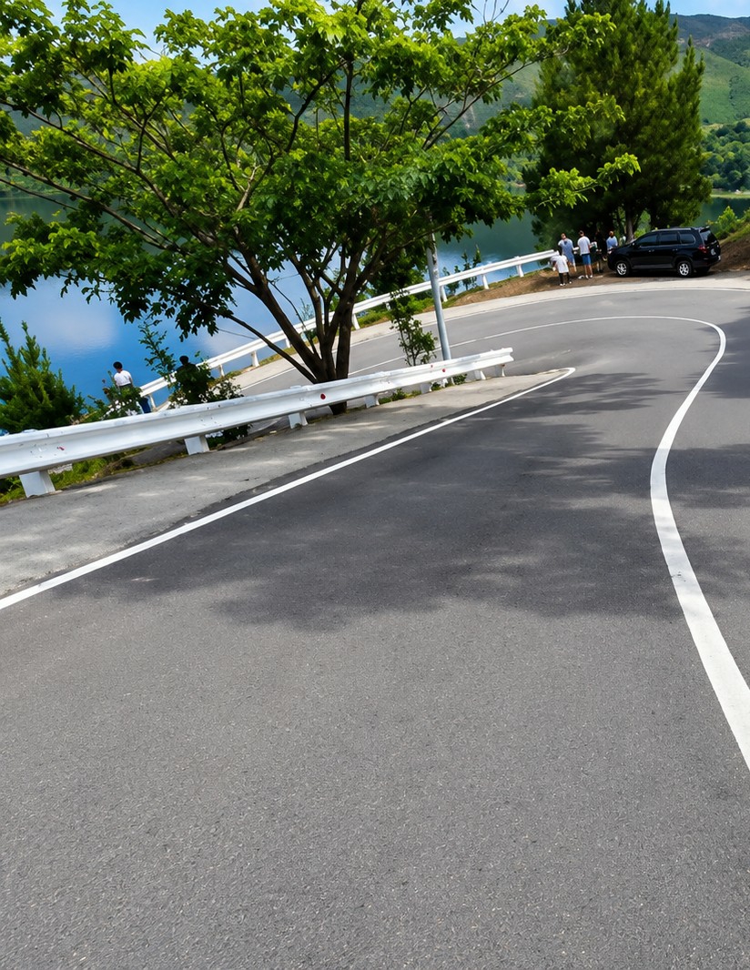 The image shows a winding paved road with a white line marking the edge. On the left side of the road, there is a body of water, likely a lake or reservoir, with a white guardrail separating it from the road. Several people are visible near the guardrail, and a dark SUV is parked on the side of the road. A large tree with green foliage is prominent in the foreground, casting shadows on the road. The background features rolling hills covered in trees, suggesting a natural or semi-rural setting. The sky is partly cloudy and light blue. This scene is located in Turpuk Sihotang, Indonesia, a location known for its natural beauty, often featuring lakes and mountainous terrain.
