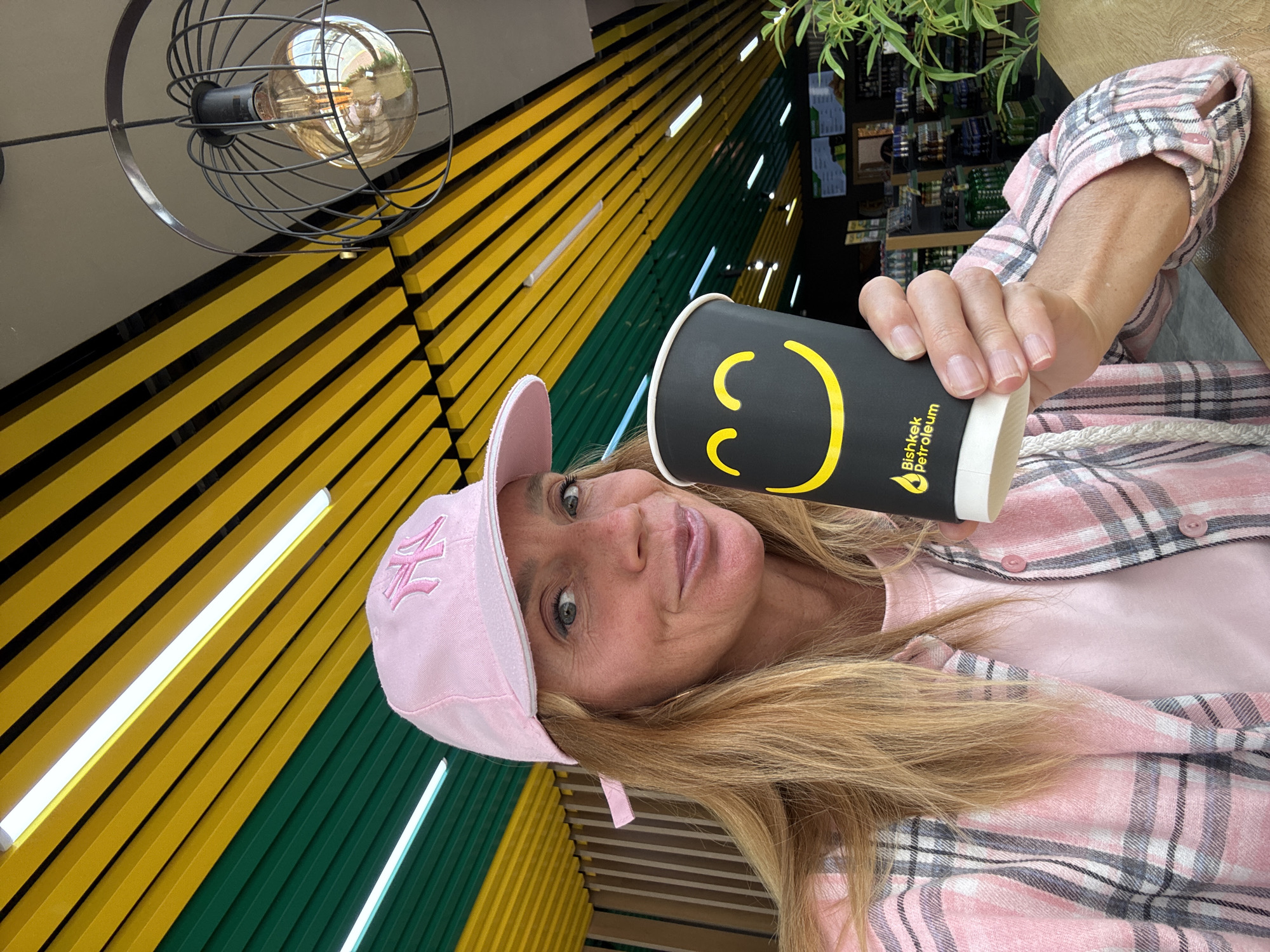 A woman is depicted indoors, looking directly at the camera while holding a black cup. She has long blonde hair and wears a light pink baseball cap with a white embroidered logo on the front, a pink plaid button-up shirt, and a plain pink t-shirt underneath. Her expression is neutral.
The black cup she holds features a prominent yellow stylized smiley face logo, along with the vertical text "Bishkek Petroleum" and a small flame or leaf icon. The background consists of a distinctive wall covered with alternating horizontal yellow and dark green slats or beams. Several long, narrow fluorescent light strips are integrated into this wall design.
In the upper portion of the frame (when reoriented to normal portrait view), an industrial-style light fixture with a black wire cage encasing an exposed Edison bulb is visible. Green foliage from a plant can be seen in the background on the right, and blurry shelves or displays are also present. The setting appears to be a modern commercial interior, possibly a cafe or convenience store, aligning with the "Bishkek Petroleum" branding on the cup and the provided context of Кашат, Kyrgyzstan. The scene is brightly lit by artificial indoor lighting.