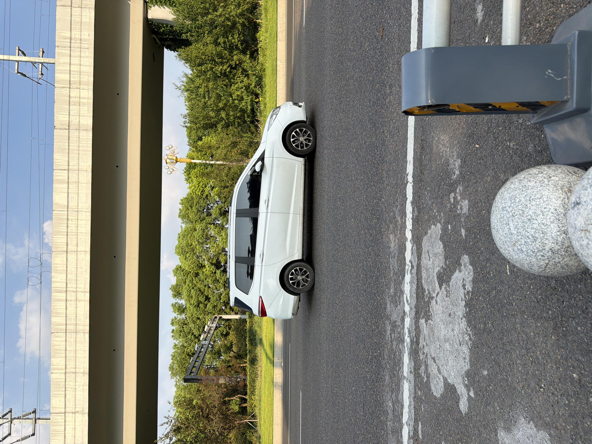 In Jiangjin, China, a white minivan is traveling on an asphalt road under a concrete overpass. The minivan, viewed from the side, is moving from left to right. The road has white lane markings. In the foreground, a grey barrier with yellow and black warning stripes and two granite spheres are visible. Behind the minivan, lush green trees form a dense backdrop. Overhead, power lines and utility poles are suspended from the overpass. The sky is blue with scattered white clouds.