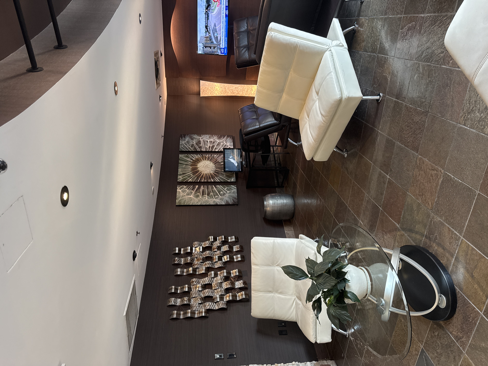 The image depicts an indoor waiting area or lounge. In the foreground, a white leather sofa is visible, complemented by a glass coffee table with a black base and silver accents. A potted plant with dark green leaves sits on the coffee table.
Further into the room, another white leather sofa is positioned to the left. The walls are dark brown, with a textured finish. A large, abstract metal wall art is mounted on the left wall. To the right, a tall, translucent floor lamp with a warm glow stands. Behind it, a flat-screen television displays a race or sporting event. Adjacent to the lamp is a black leather armchair. In the background, a desk with a computer monitor and a black chair is set up. The flooring is made of large, dark square tiles with a textured surface.
The image appears to be taken in a commercial or hospitality setting. Without further information, the specific location within North Vancouver, Canada, cannot be determined from the visual evidence alone.