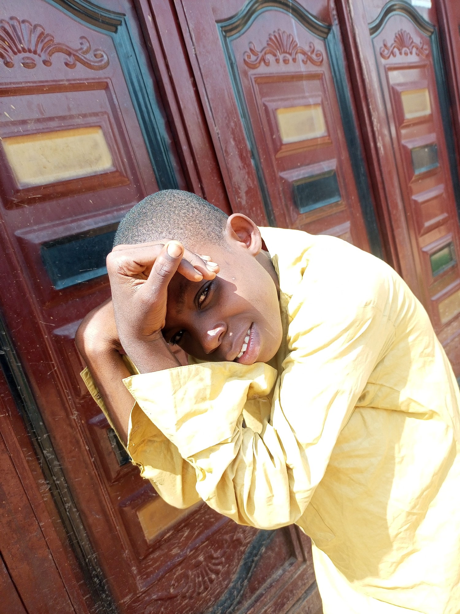 A young person with dark skin and short hair is leaning against a wooden door. They are wearing a light yellow collared shirt. Their right arm is bent at the elbow, and their hand is resting on their forehead, partially obscuring their face. The person is looking towards the camera and smiling. The door is dark red with decorative carvings and rectangular panels. The lighting suggests it is daytime, with sunlight casting shadows.