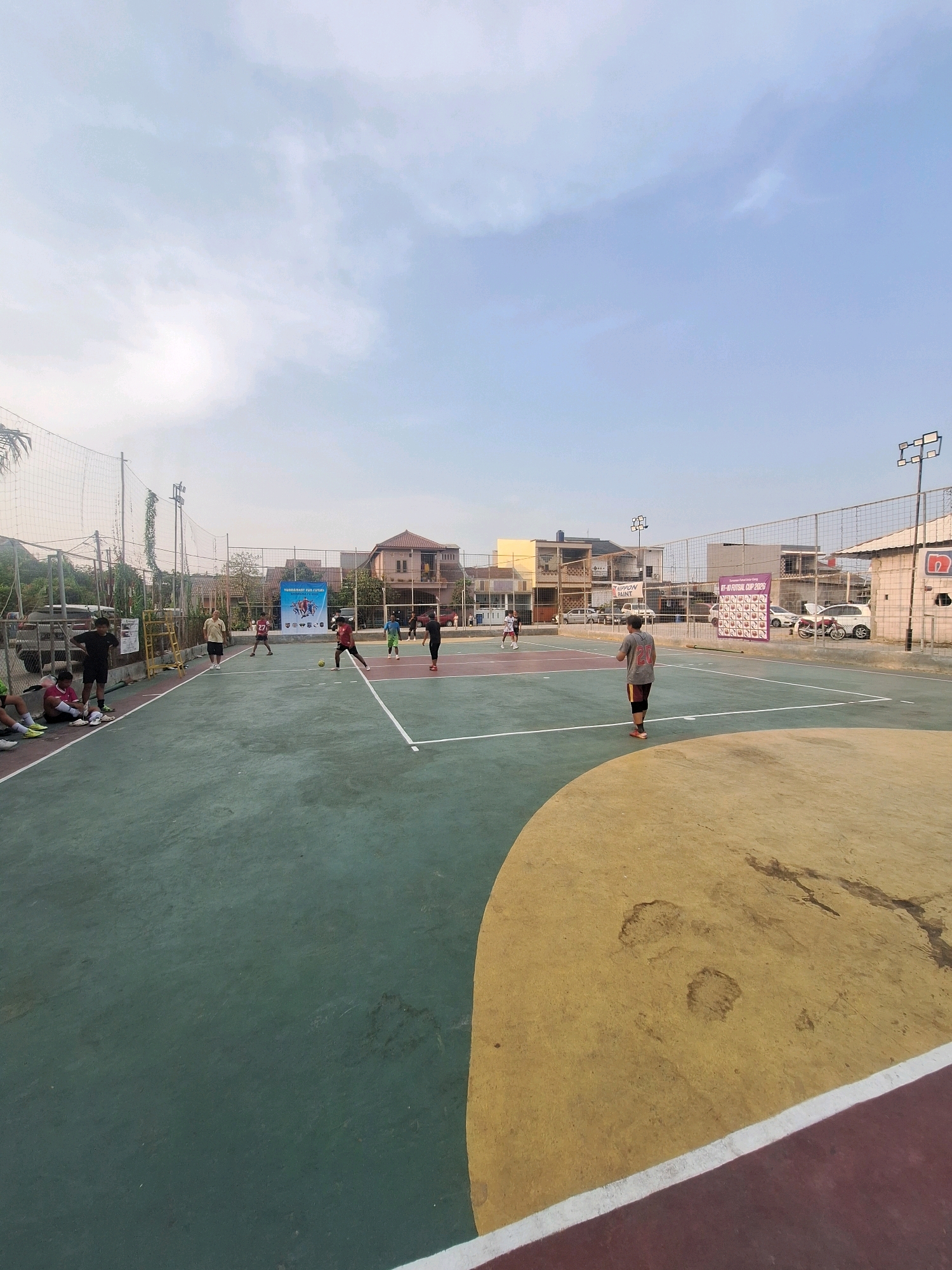 The image depicts an outdoor futsal court in Babelan, Indonesia, on a clear day with a light blue sky. Several individuals are present on and around the court. On the court, a group of people are engaged in a futsal match, with some actively playing and others observing from the sidelines. A banner promoting a "FUTSAL CUP 2023" event is visible in the background, along with buildings and parked vehicles. The court itself is marked with white lines and features different colored sections of pavement. The overall scene suggests a community sporting event or recreational activity taking place in an urban environment.