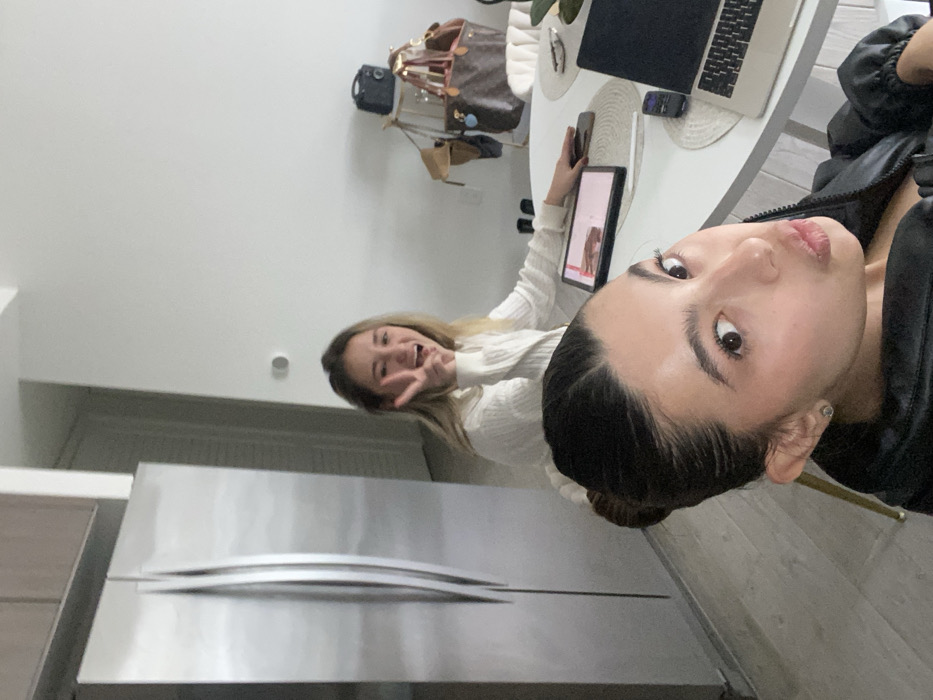 Two women pose in a selfie. One woman wearing a black jacket is close to the camera, with a slight pouting expression. Behind her to the left, another woman in a white sweater smiles and makes a peace sign. A stainless steel refrigerator is visible at the bottom. A white table with a laptop and a tablet sits behind the women. A handbag hangs on the wall.