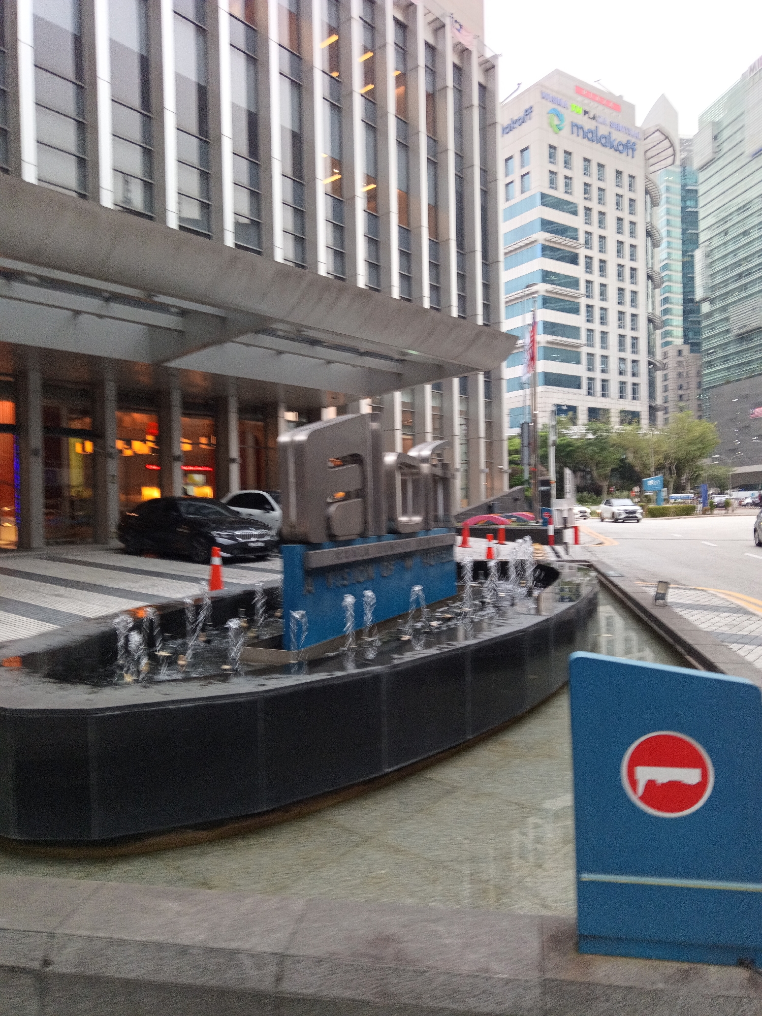 The image depicts an urban scene in Kuala Lumpur, Malaysia, likely a commercial or business district given the modern architecture. In the foreground, a dark, rectangular water feature with several spouts is visible. A blue sign with a red circle and a white horizontal bar, indicating a prohibition or restriction, stands to the right of the water feature.
Behind the water feature, a black sedan is parked. Adjacent to it, a white car is also visible. The entrance to a building, characterized by a gray canopy and a series of columns, is to the left. The building facade features numerous vertical windows.
In the background, several tall, contemporary buildings dominate the skyline. One prominent building displays the "Malakoff" logo and text. The overall environment suggests a well-developed urban setting with a focus on business and corporate presence. The lighting indicates daytime, possibly overcast, as there are no strong shadows. The image appears to be captured from a street-level perspective.