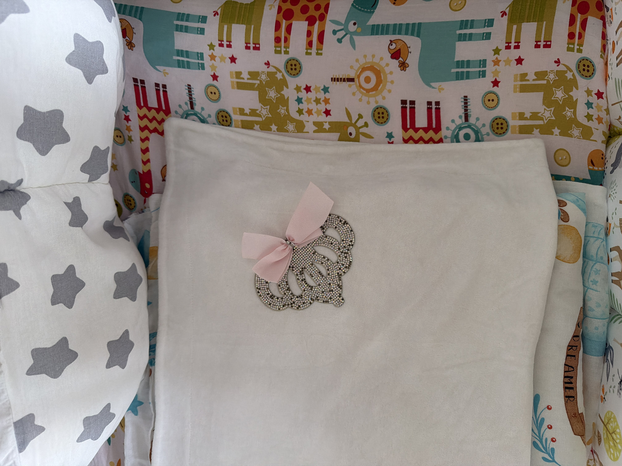 A close-up, overhead shot shows a white crib with a patterned sheet featuring giraffes, stars, and buttons. A white blanket is folded over the side of the crib. A decorative crown-shaped embellishment with a pink bow is placed on the white blanket. The image is taken indoors, likely in a nursery. There is no information available about the location in Moscow, Russia.