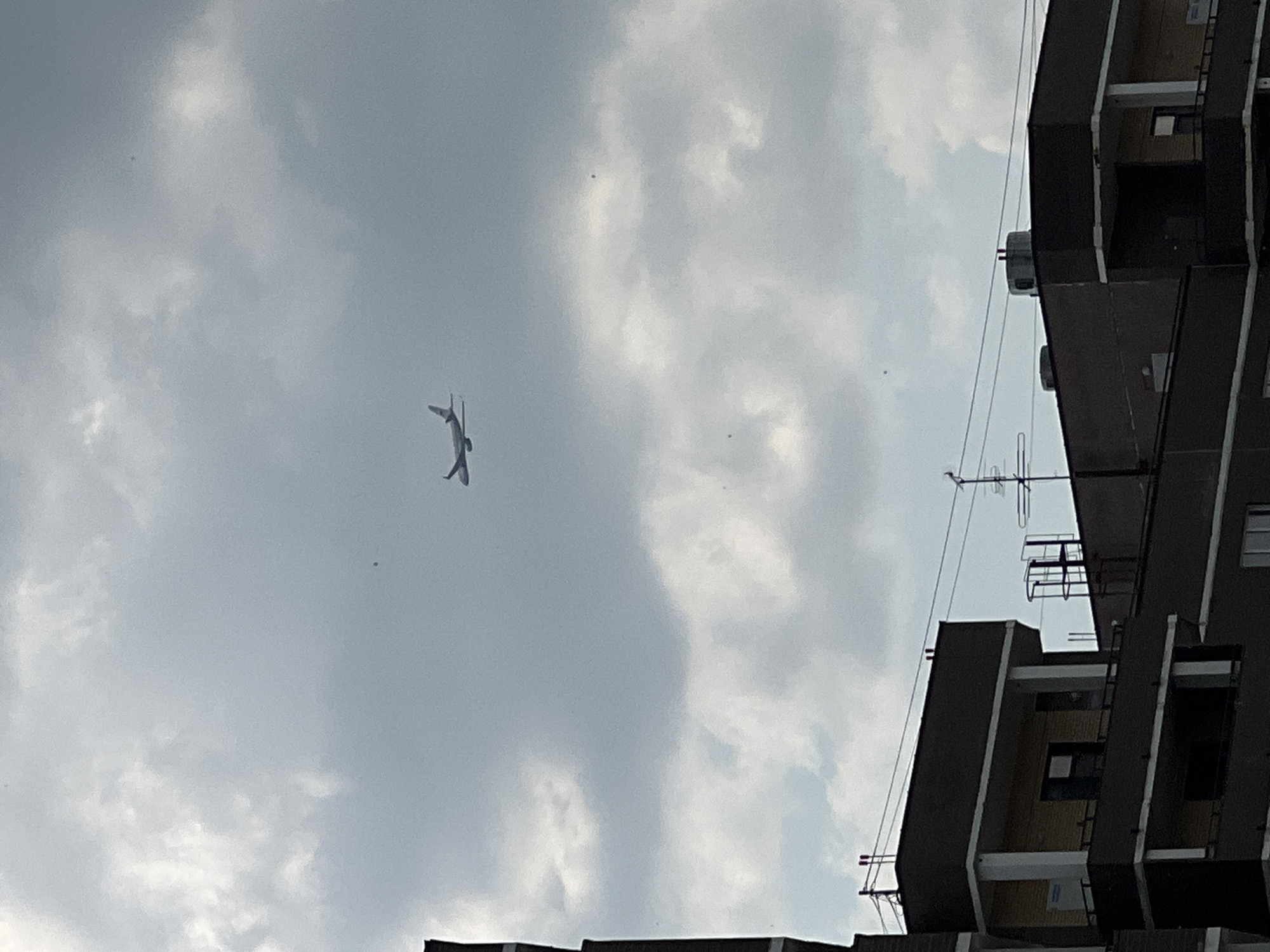 The image shows an airplane flying in a cloudy sky above residential buildings. The buildings appear to be apartment complexes with multiple stories. The sky is overcast with various shades of gray and white clouds. The airplane is visible in the upper portion of the frame, silhouetted against the sky. The scene is consistent with an urban environment, possibly taken from a residential window or balcony. The specific location in Novosibirsk, Russia, cannot be determined from the visual information alone.