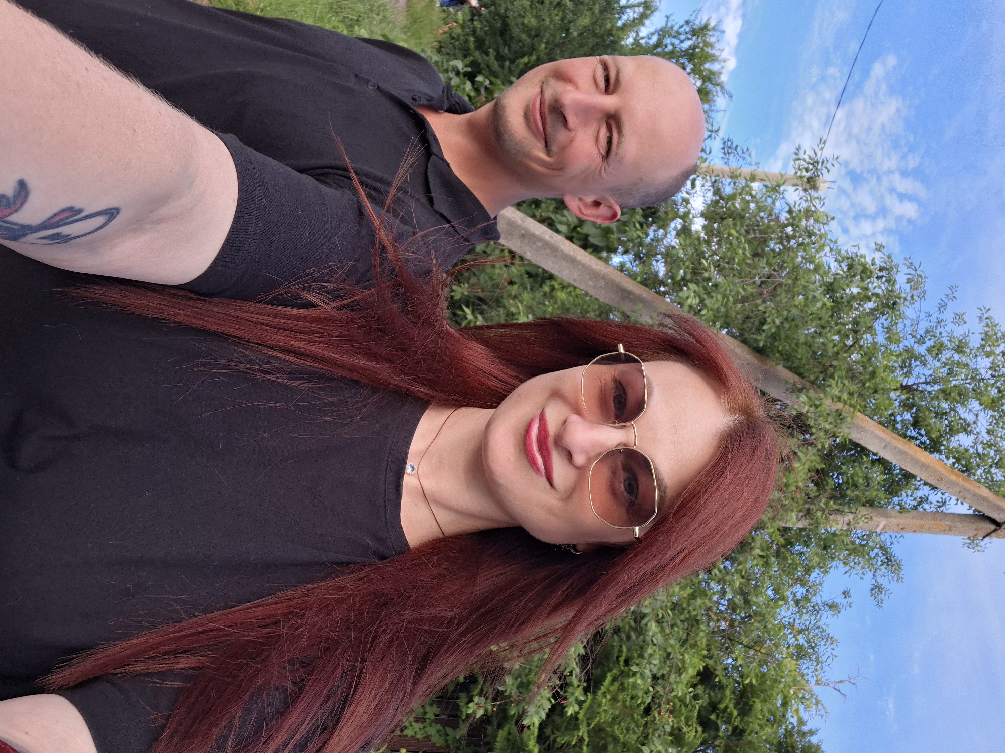 A man and a woman are posing for a selfie outdoors in Забалоцкі сельскі Савет, Belarus, with lush green trees and a blue sky with white clouds in the background. The man, balding with fair skin, is wearing a black polo shirt and smiling. The woman, with long, reddish-brown hair and sunglasses, is wearing a black t-shirt and a small pendant necklace. Her left arm, visible in the foreground, has a tattoo. A concrete pole and some fencing are also visible in the background. The overall impression is a casual outdoor portrait.