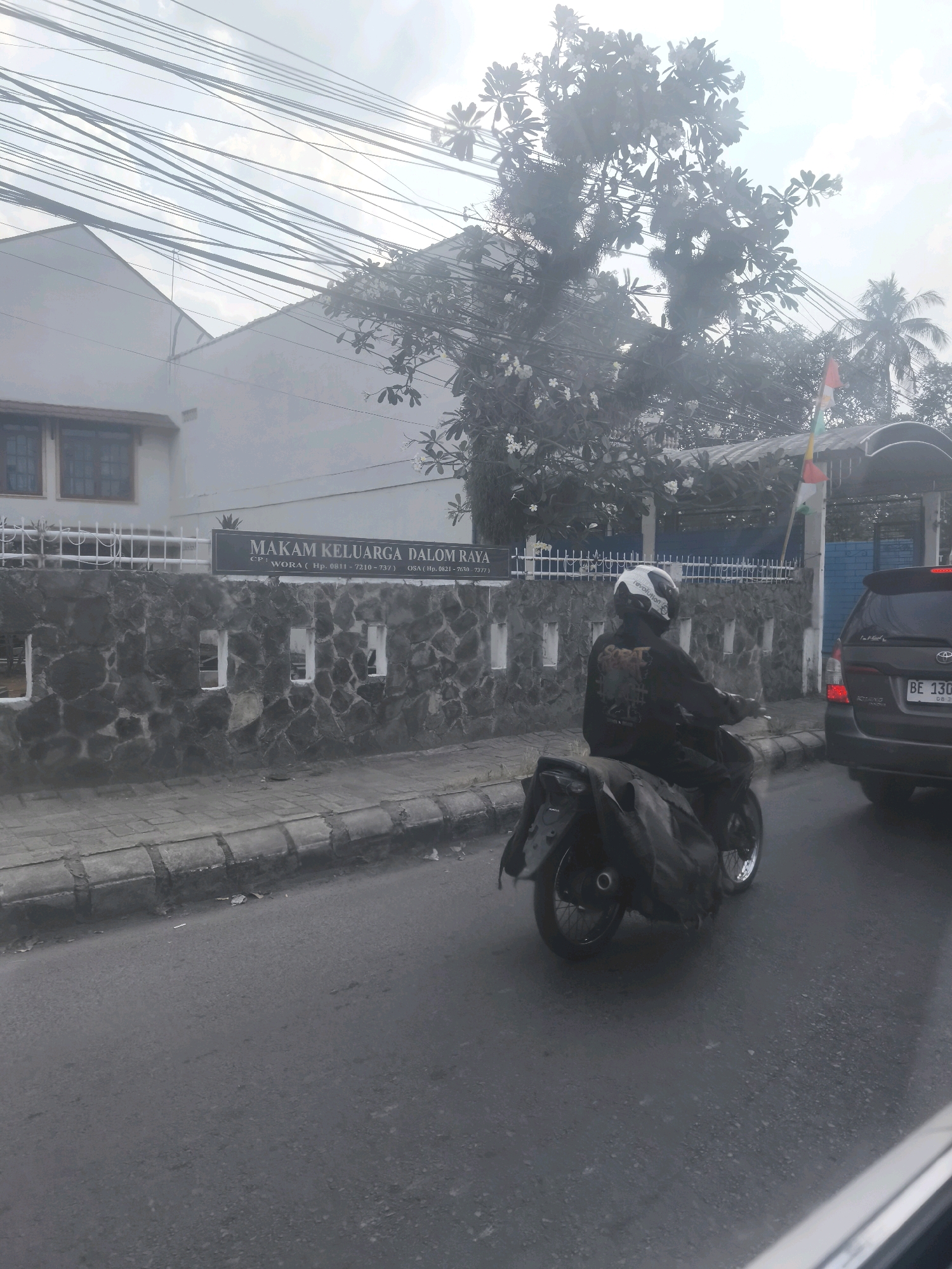 A motorcycle rider wearing a black jacket and white helmet is traveling down a street in Bandar Lampung, Indonesia. The rider is on a black motorcycle with bags attached to the back. To the right of the motorcycle, a dark-colored car with a license plate reading "BE 130..." is also on the road. The street is paved and has a sidewalk. Behind the motorcycle, there is a stone wall with a sign that reads "MAKAM KELUARGA DALOM RAYA". Beyond the wall, there is a building with windows and a tree with white flowers. The sky is overcast with power lines crisscrossing the image.