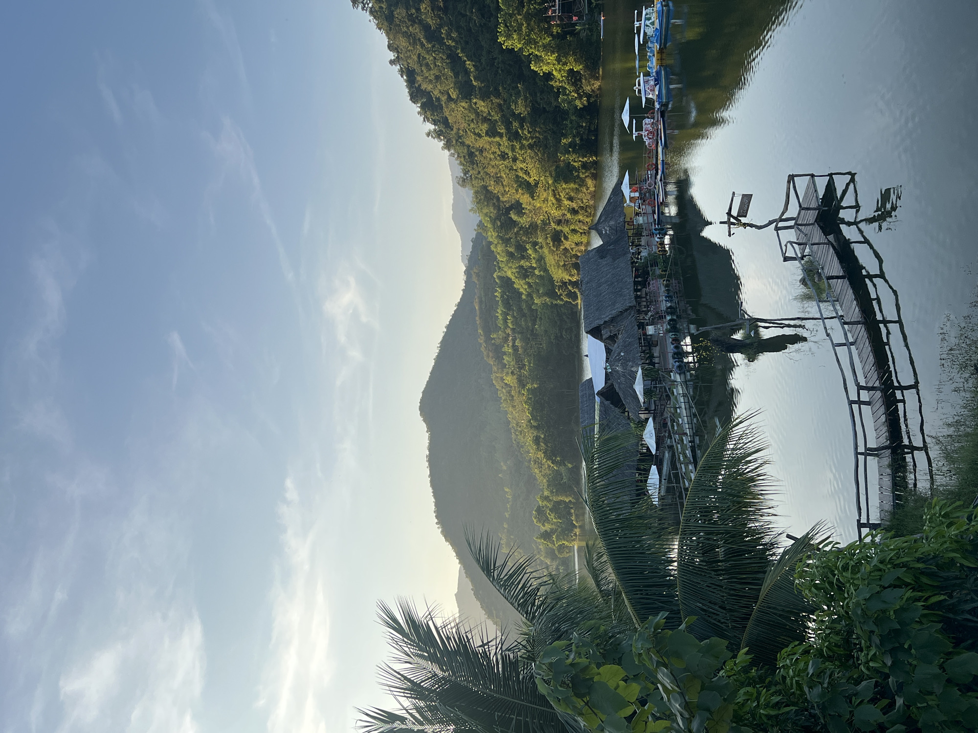 This image captures a panoramic view of **Hồ Ba Hô (Ba Ho Lake)**, approximately 25-30 km north of Nha Trang, Khánh Hòa Province, Vietnam. The scene features a tranquil lake surrounded by multiple layers of verdant, forested mountains under a clear to partly cloudy light-blue sky with faint cirrus clouds. Soft, warm sunlight illuminates the distant mountain peaks and upper slopes, suggesting either early morning or late afternoon.
In the immediate foreground, on the left, large, dark green palm fronds and other dense tropical foliage partially obscure the view. On the lake's calm surface in the midground, a cluster of floating structures with dark, conical thatched roofs is present, connected by wooden walkways. Several white sun umbrellas are positioned among these structures. To the right of these floating buildings, a dock area is visible, where multiple brightly colored pedal boats (blue, yellow, white) are moored. A number of red and white lifebuoys hang on the dock's perimeter.
In the lower right foreground, a rustic wooden boardwalk or pier extends from the vegetated bank into the lake, curving slightly to the right. A wooden signpost with an illegible sign is affixed at the end of this pier. The entire boardwalk and signpost are clearly reflected in the still water. No individuals are visible in the scene. The lake water is dark, reflecting the sky and surrounding landscape, creating a mirror-like effect.