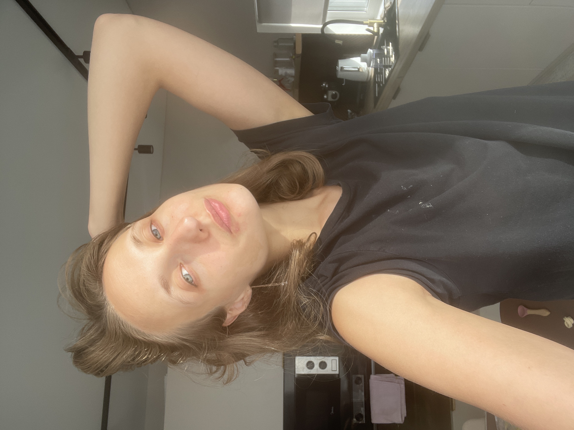 A young woman with light brown hair and blue eyes is taking a selfie in a kitchen. She is wearing a black t-shirt and has her right arm raised behind her head. The background shows kitchen cabinets, a microwave, and a countertop with a sink and a kettle. Natural light is coming in from a window on the right side of the image. The image was taken in the city of Podshivalovo, Russia.