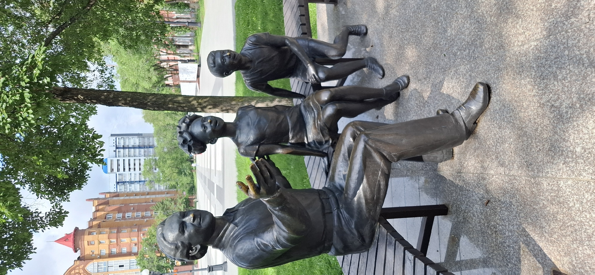 A bronze sculpture depicts three figures seated on a park bench in Blagoveshchensk, Russia. In the foreground, an adult male figure on the left is gesturing with his hands. To his right, a young girl with an elaborate hairstyle sits with her hands in her lap, followed by a young boy on the far right, also seated. All three figures have what appear to be small, gold-colored objects on their noses, possibly representing something specific to the sculpture's theme or a local tradition. The figures are rendered with a realistic, yet slightly stylized, appearance. The park bench is made of wooden slats and has dark metal legs. The background features a paved walkway, green grass, trees, and several modern buildings, including a brick building with a red turret and taller, lighter-colored apartment buildings. The lighting suggests a daytime scene under a partly cloudy sky, casting shadows on the ground.